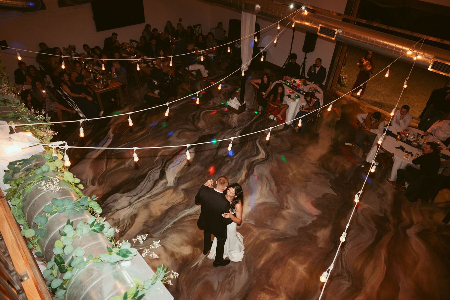 Couples dancing under romantic string lights at an indoor wedding reception venue.