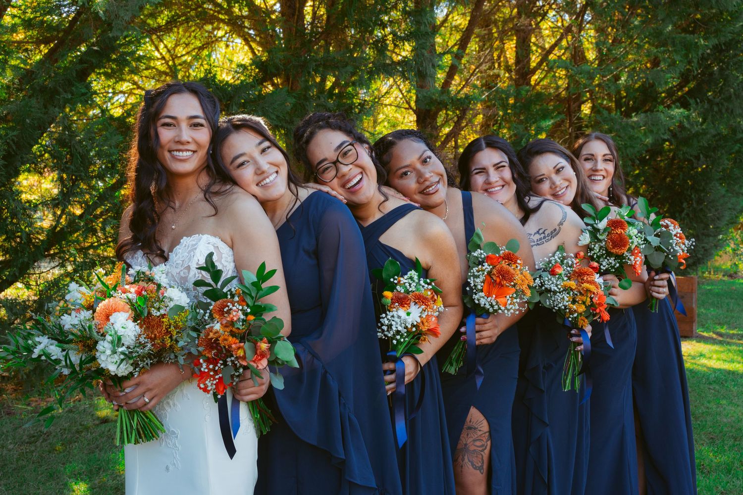 Wedding group stands in line wearing navy dresses and holding orange bouquets in outdoor garden setting.