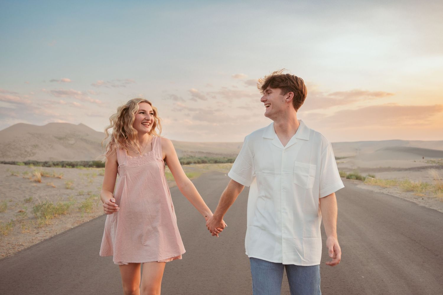 A couple in casual attire walks together on a remote desert highway at sunset.