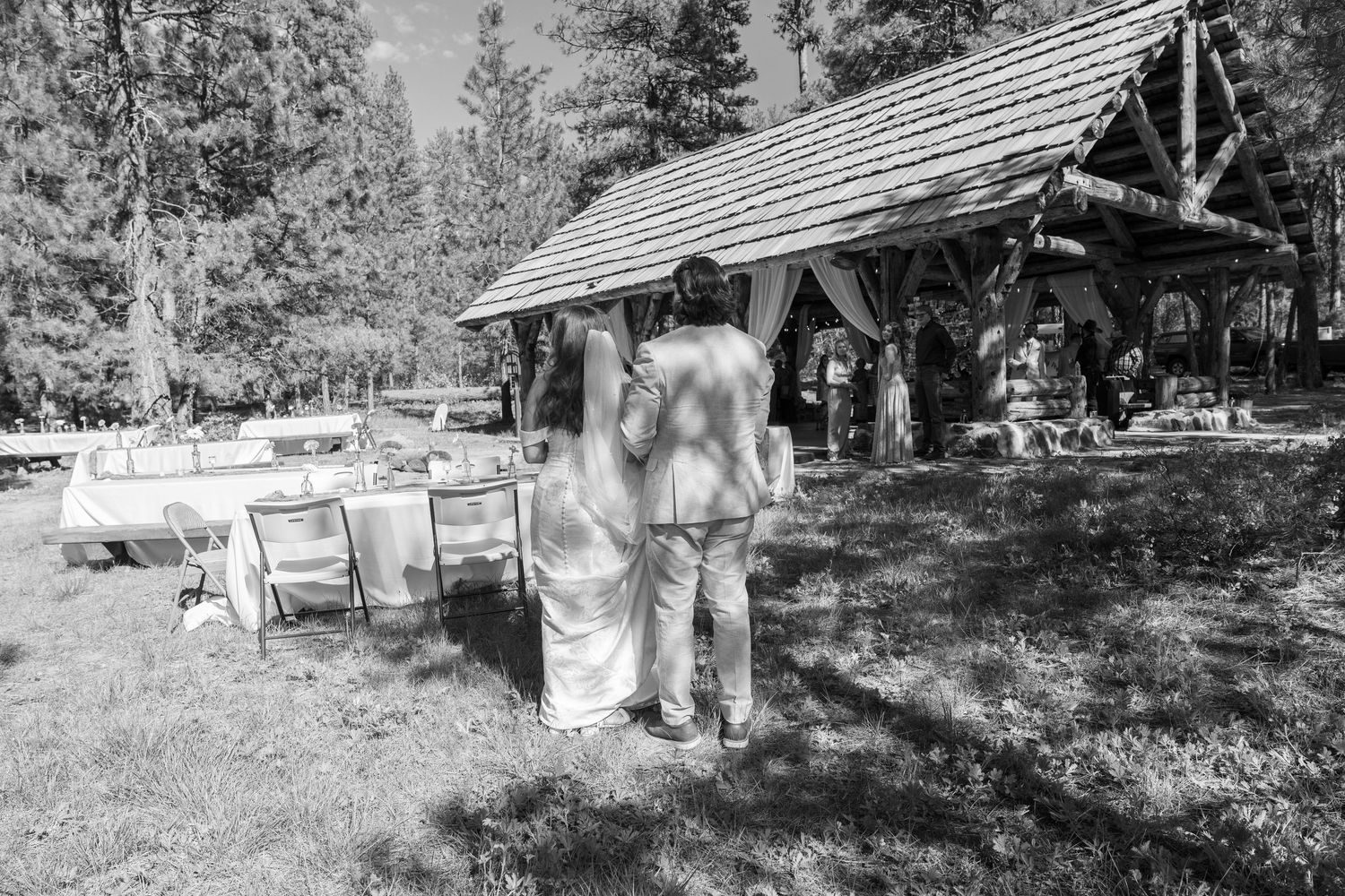 A black and white photo of a rustic outdoor wedding reception area with wooden pavilion.