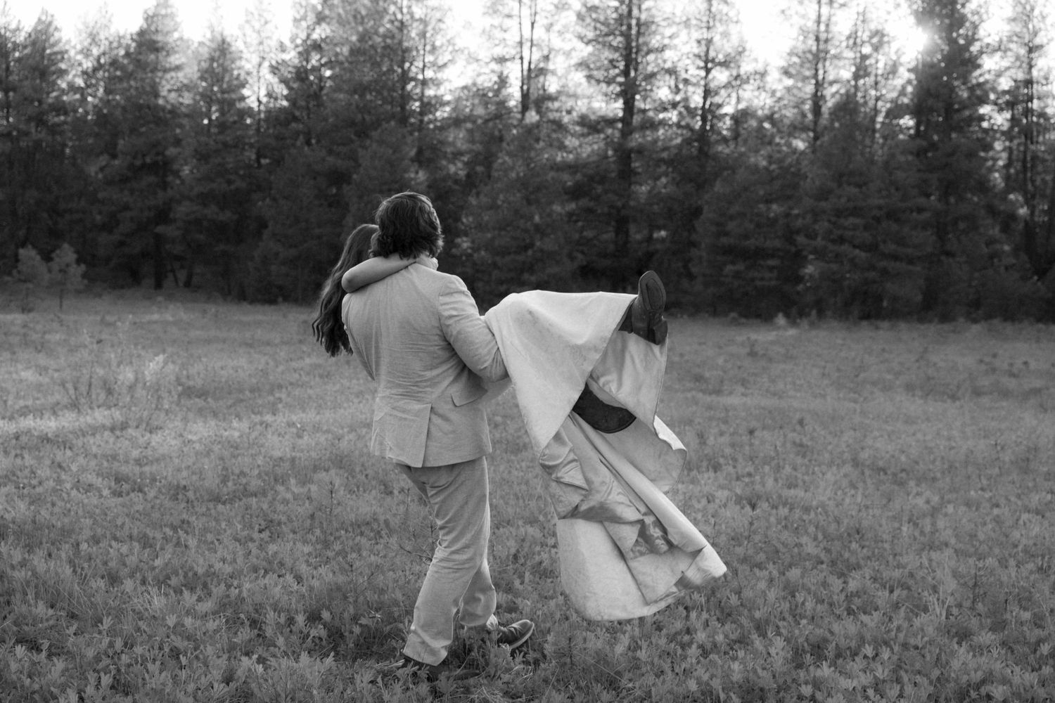 Monochrome image of a couple twirling in a meadow with a forest backdrop.
