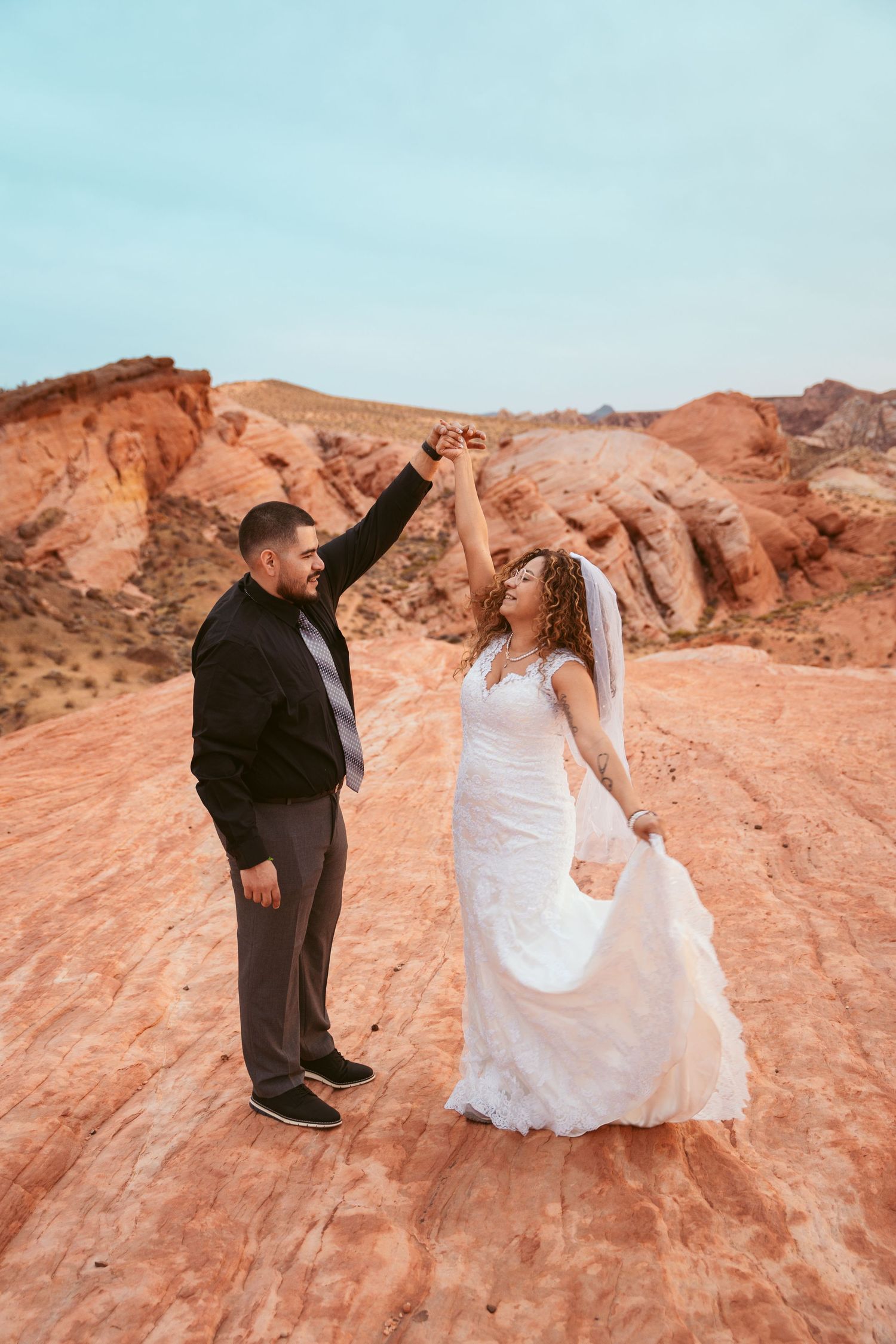 A wedding couple poses playfully on red rock formations in a desert landscape at sunset.