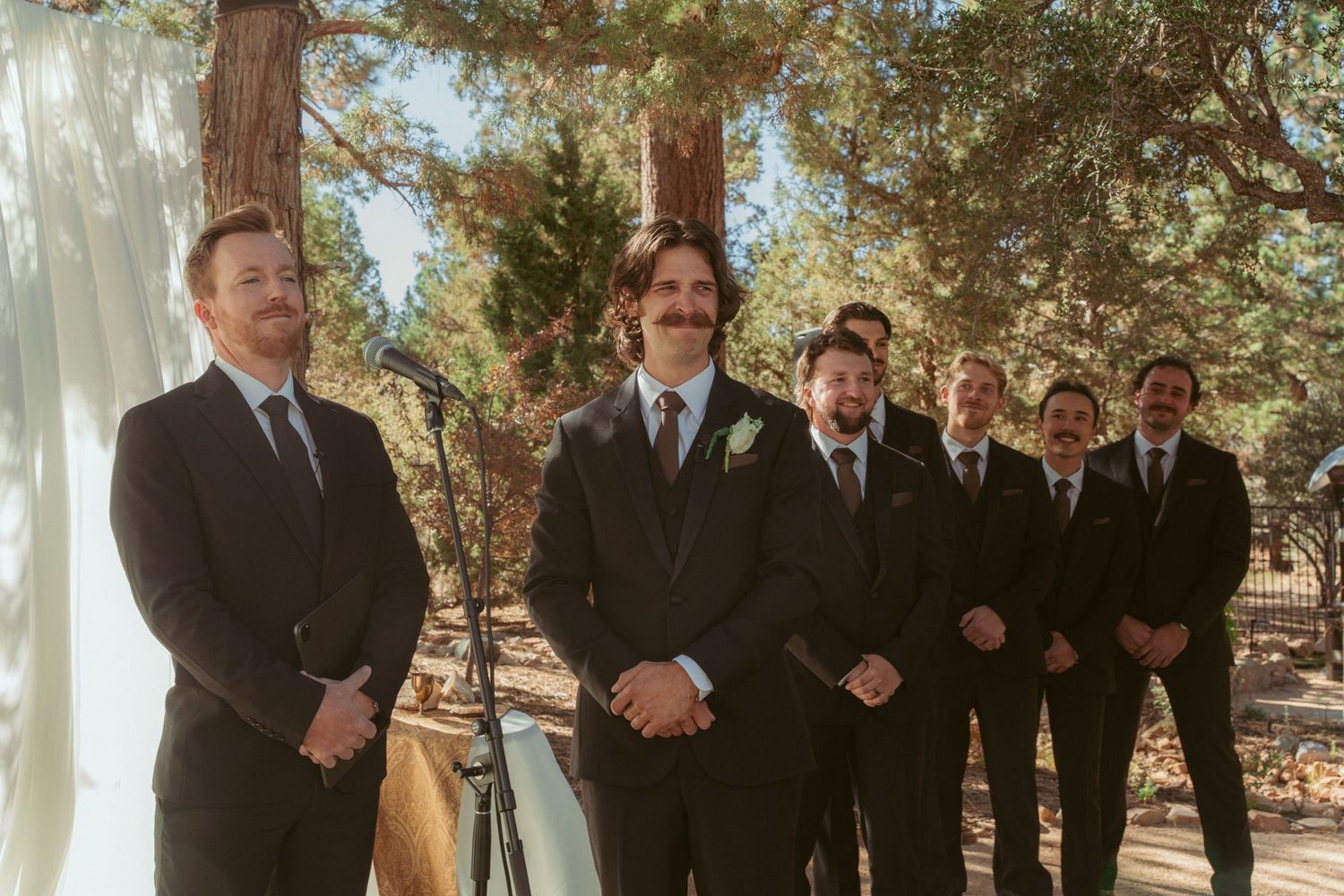 Wedding party members lined up during an outdoor ceremony beneath tall trees.