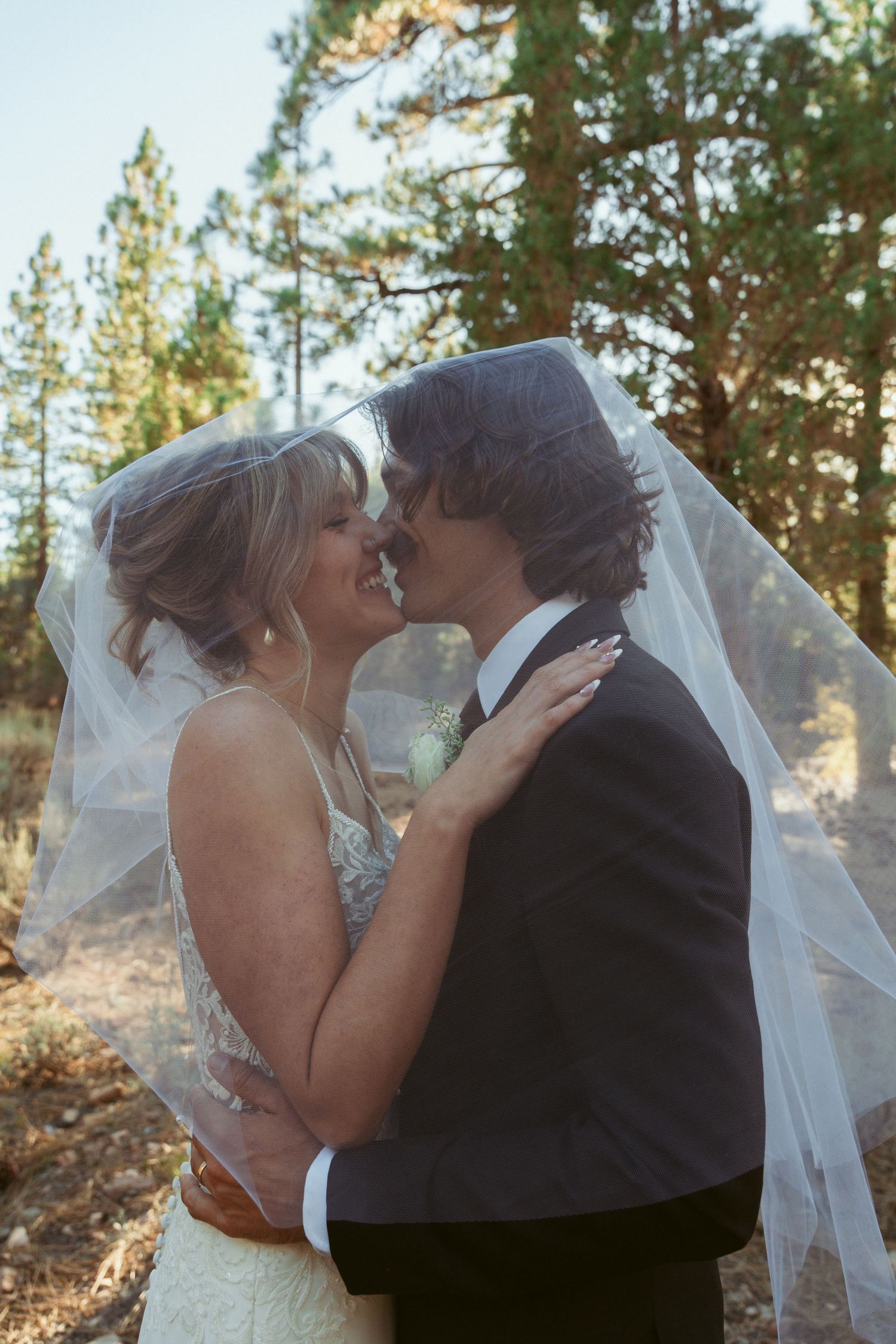 A romantic wedding couple embraces under a flowing veil in a forest setting.