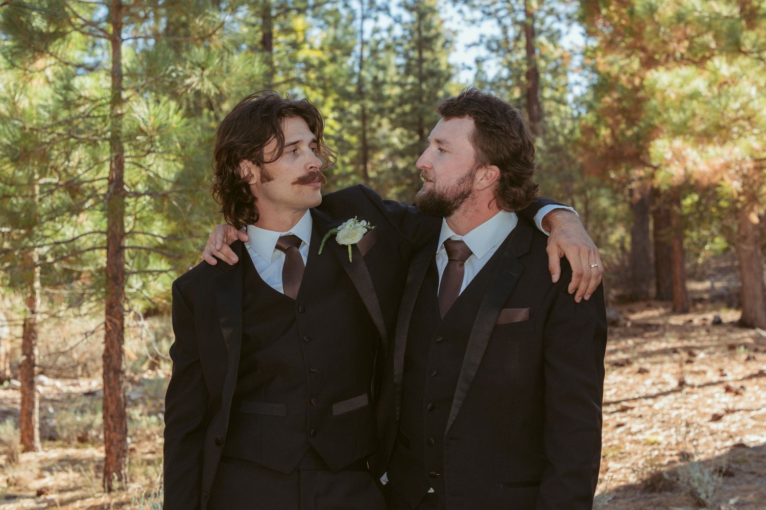 Two people in dark suits share a tender moment among pine trees.