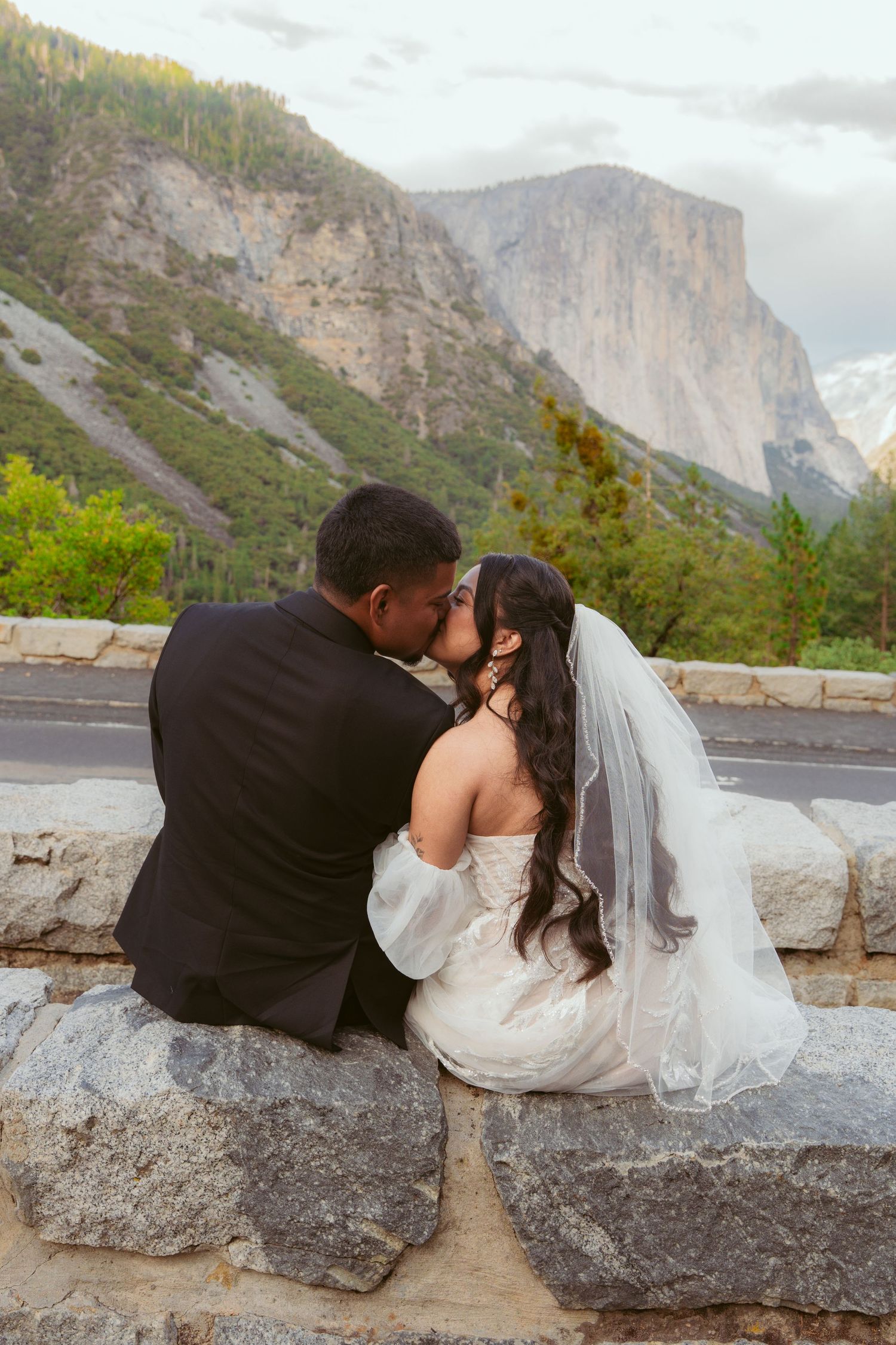 Couple in wedding attire sharing romantic moments on stone wall with mountain vista.