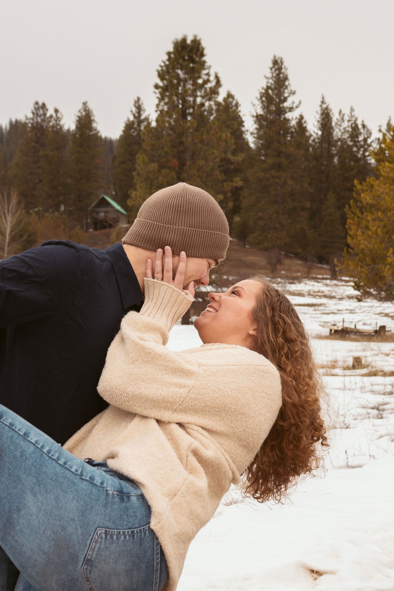 Intimate winter photoshoot with couple in cream sweater and brown beanie against snowy forest backdrop.