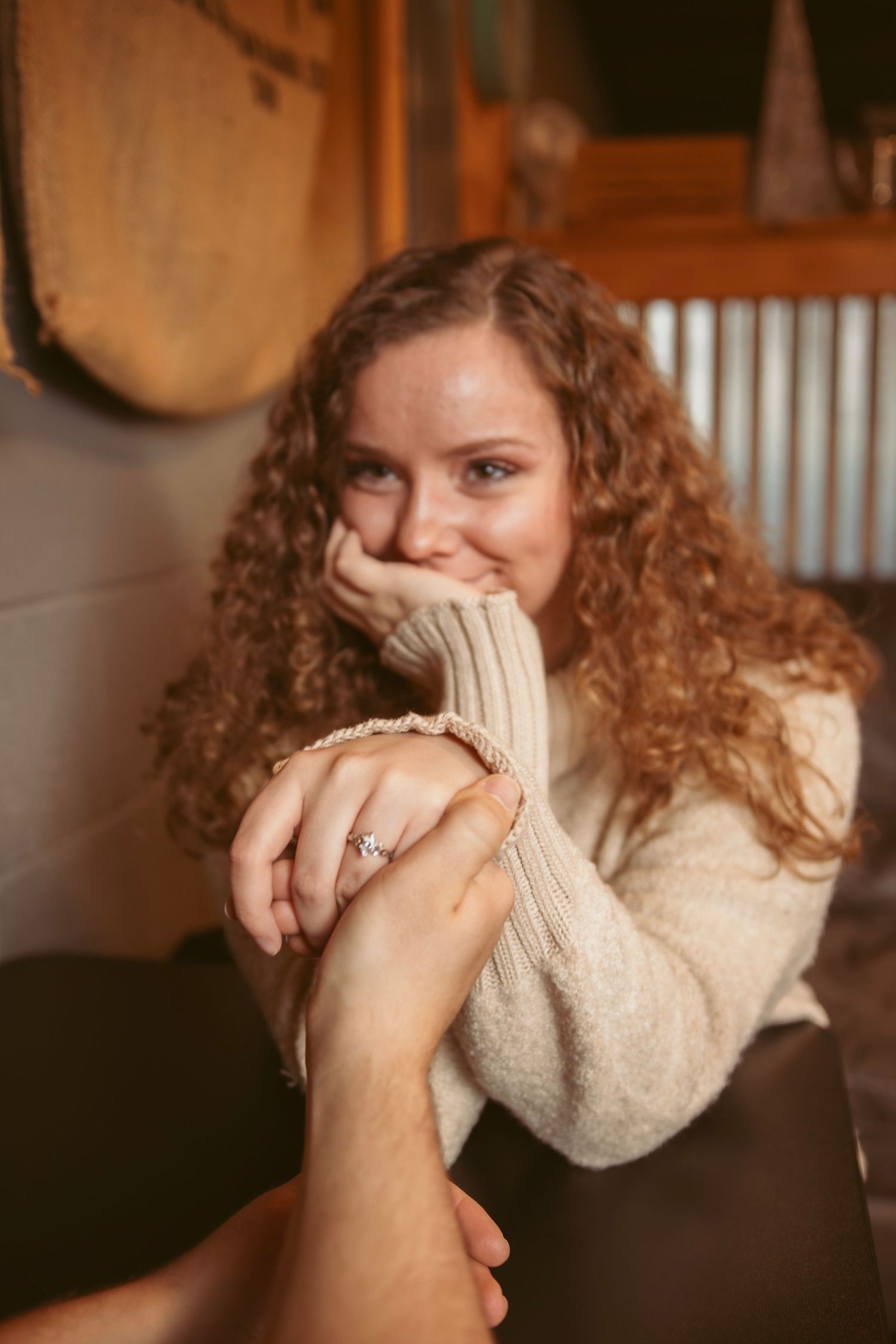 Person with curly hair expressing joy while showing engagement ring in cafe.