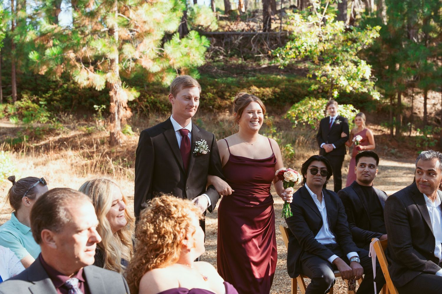 Wedding processional taking place outdoors among tall pine trees.