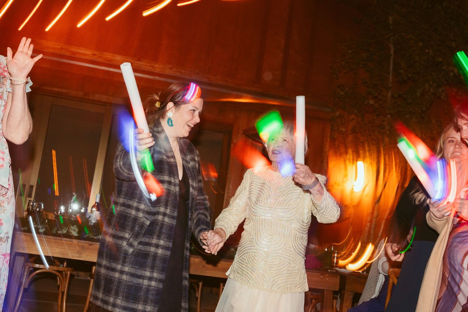 Wedding party dancing with colorful glow sticks under string lights at night.