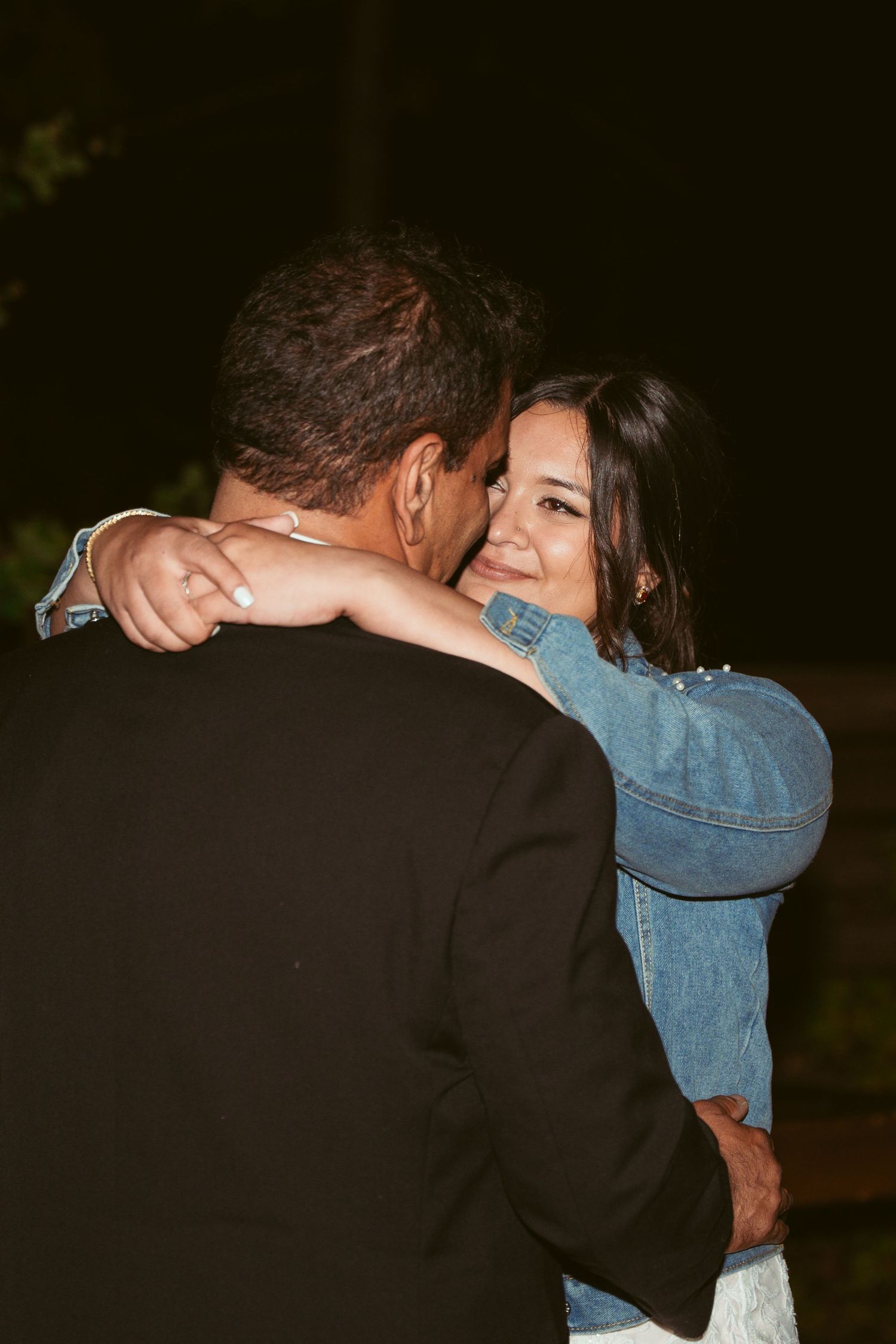 A couple embraces during their wedding dance wearing a black suit and denim jacket.