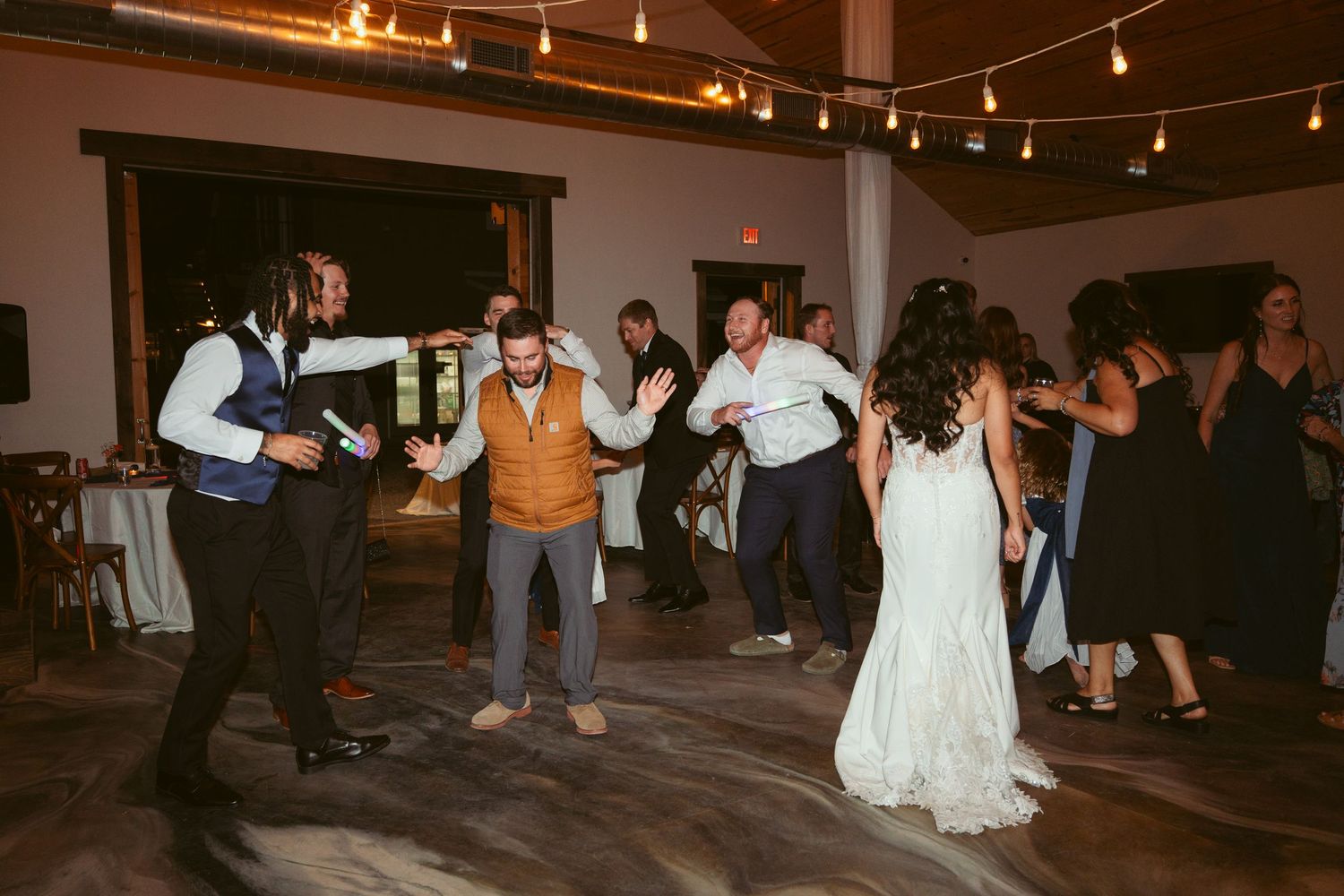 Wedding guests dancing in an indoor venue decorated with string lights and white draping.