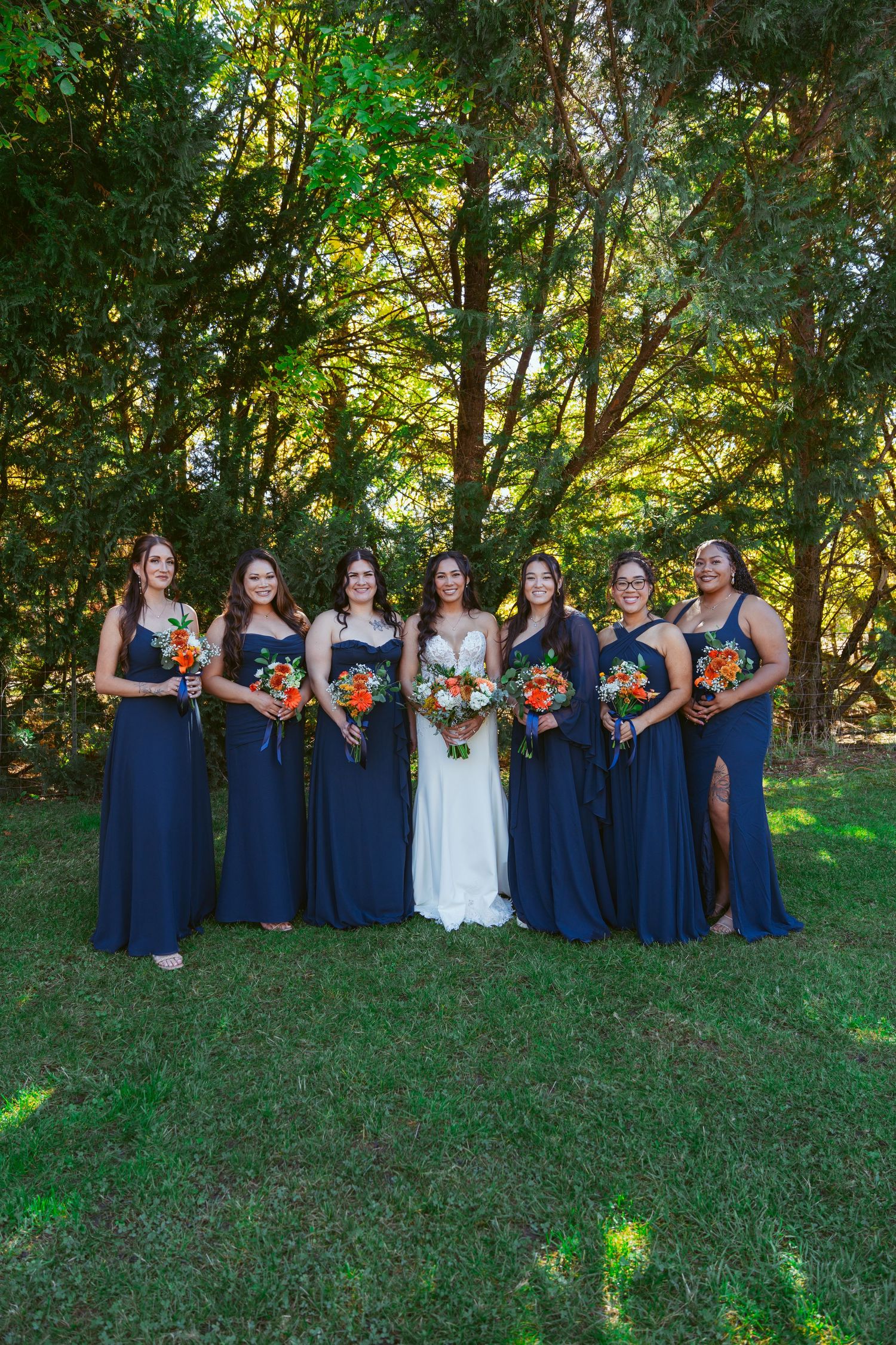 Bridal party in navy blue dresses poses with bouquets against lush green forest backdrop.