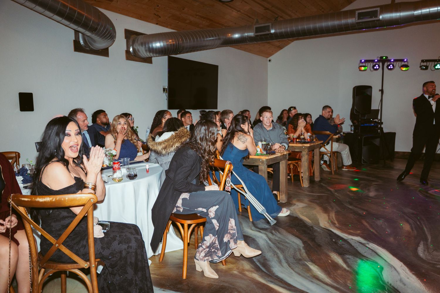 Wedding guests seated at tables in a rustic reception venue with wooden crossback chairs.