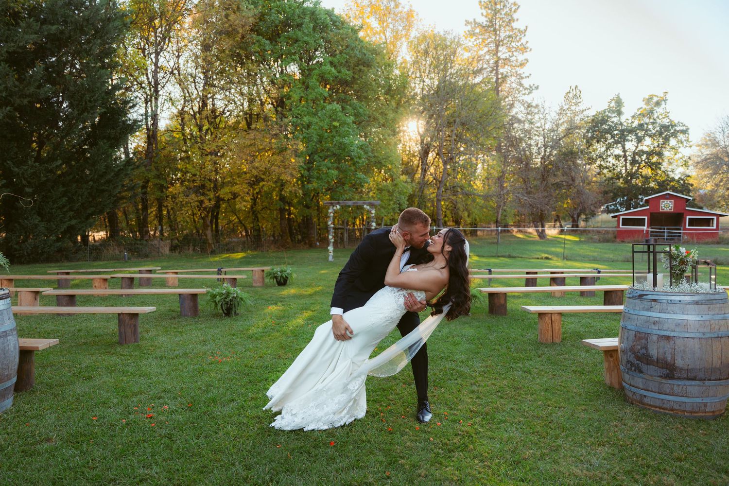 Romantic dip pose between newlyweds on lawn with wooden benches and barrels in background.