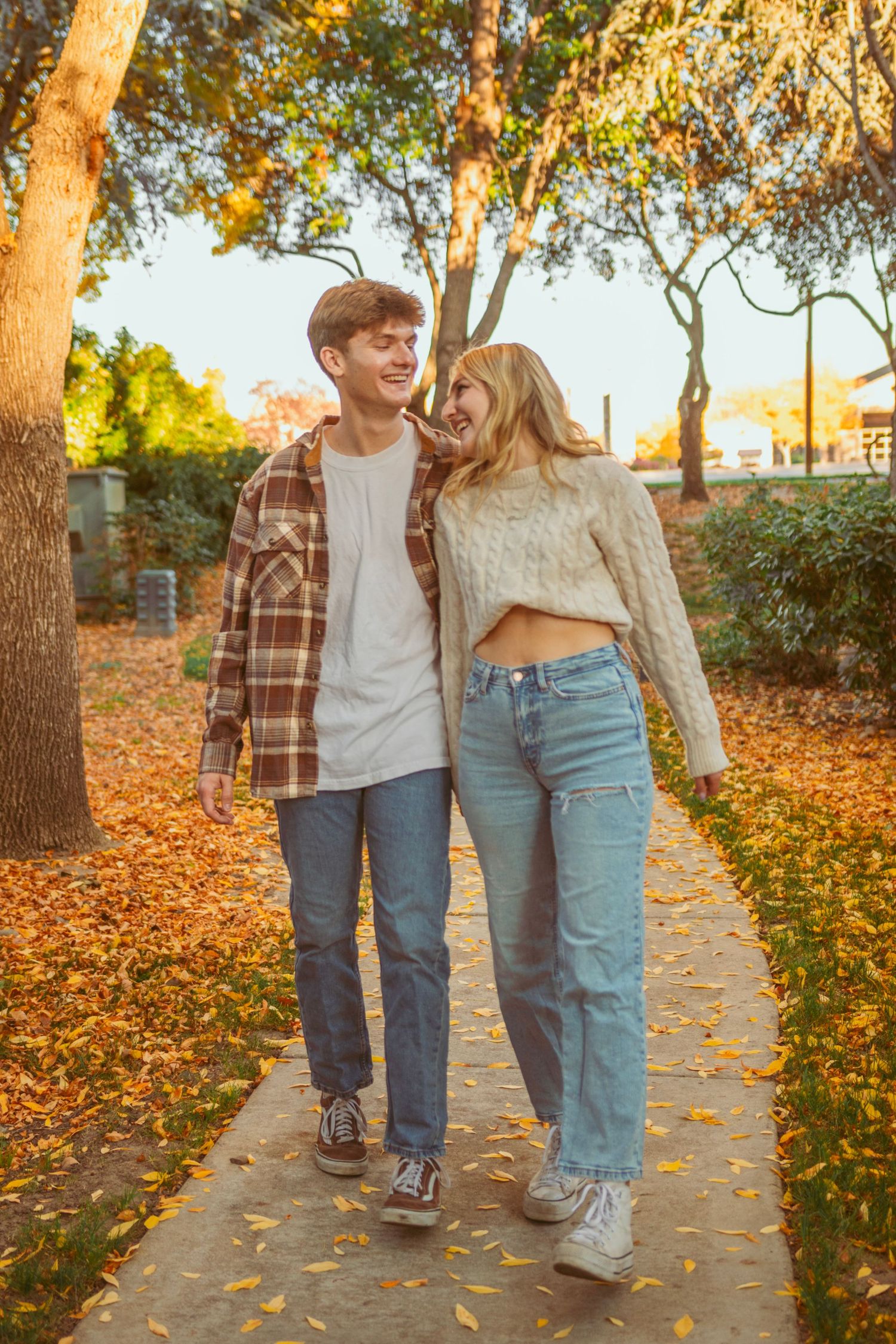 A couple walks together on a leaf-covered path beneath autumn trees.