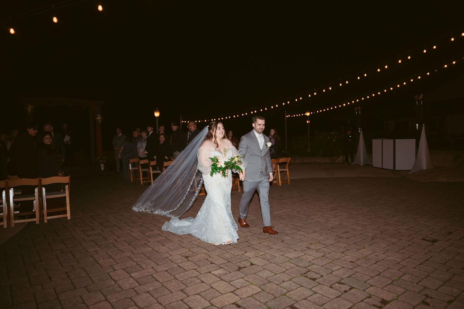 Couple walking together under string lights at their evening outdoor wedding.