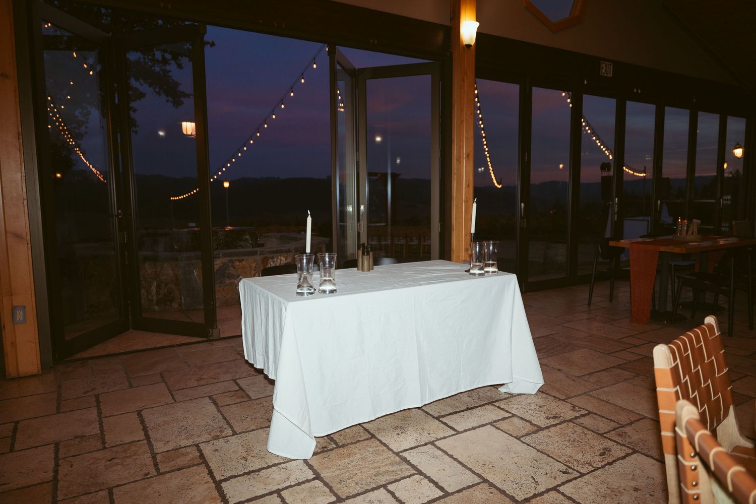 Evening reception setup with white tablecloth and string lights visible through windows.