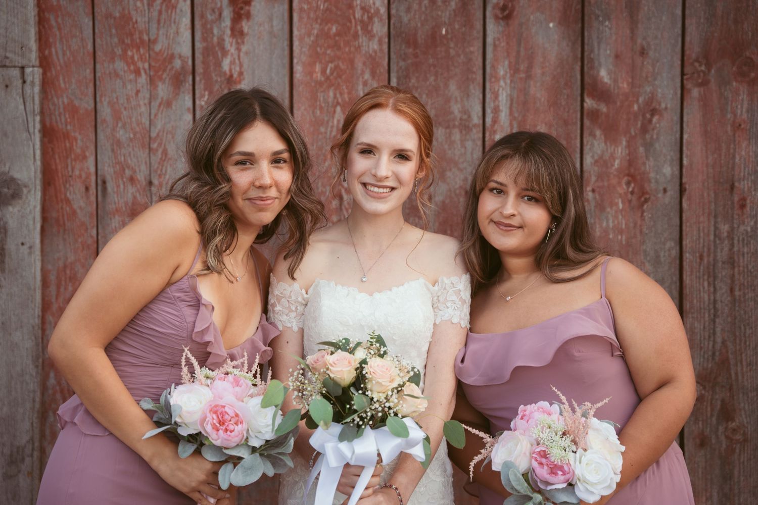 Bridal party wearing dusty rose gowns pose with bouquets against rustic wooden backdrop.