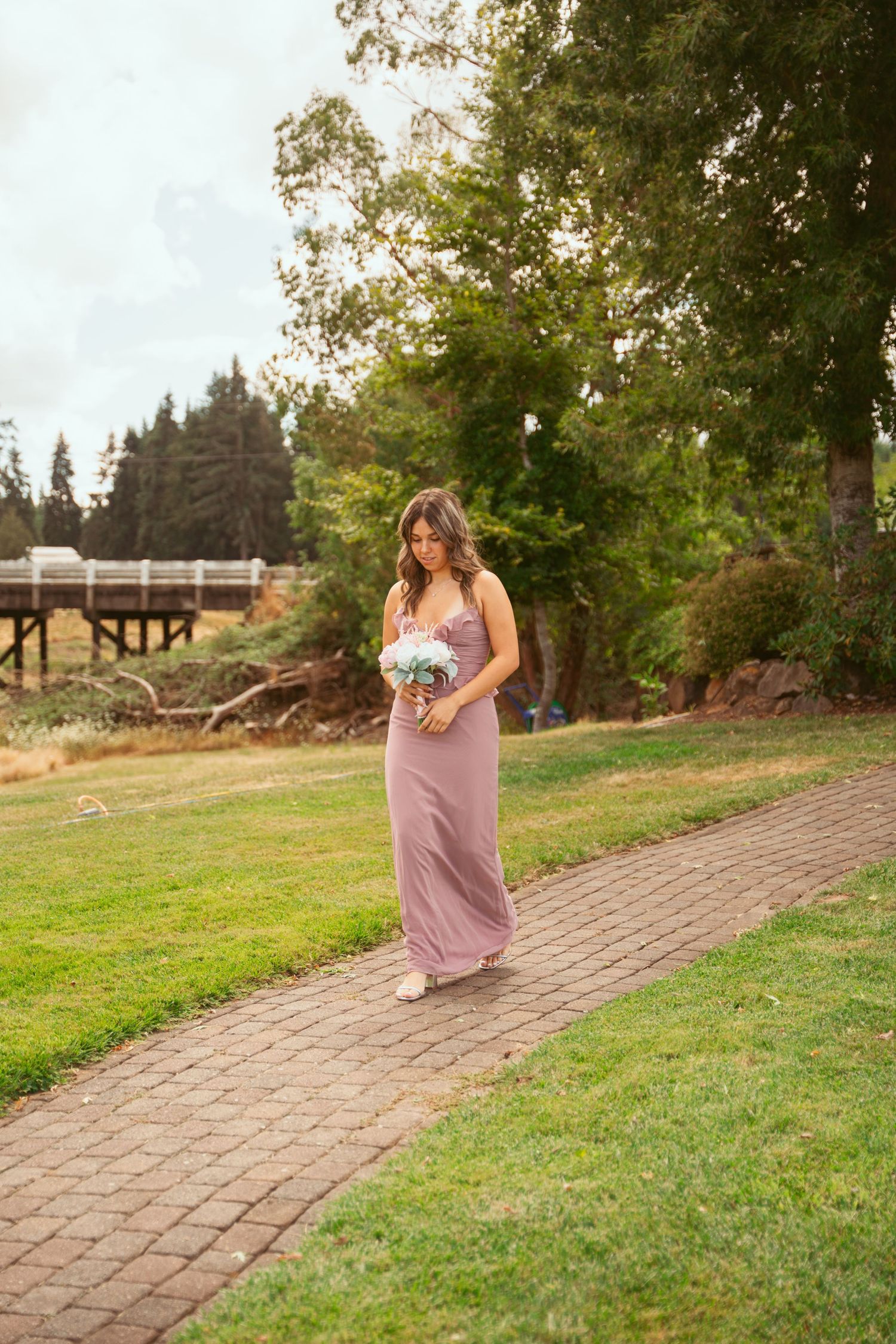 A person in a mauve dress holds flowers while walking along a curved brick pathway outdoors.