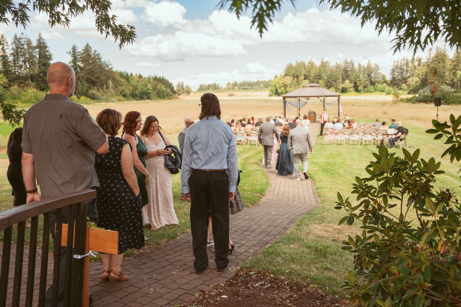 Wedding guests watch outdoor ceremony from a brick pathway overlooking a grassy field.