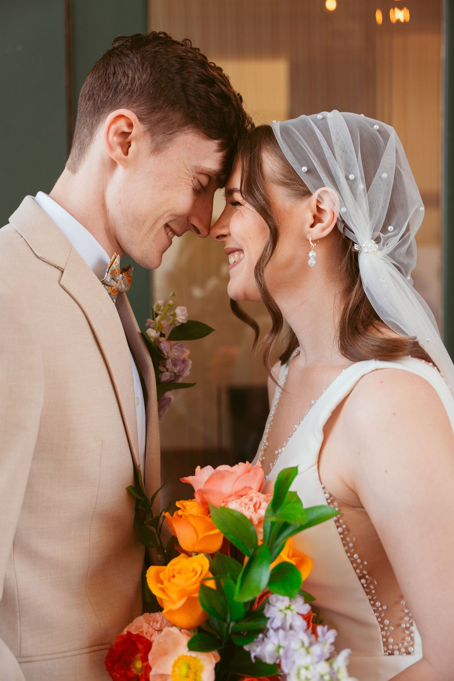 A tender moment between newlyweds with a vibrant orange and pink bouquet against a soft interior backdrop.