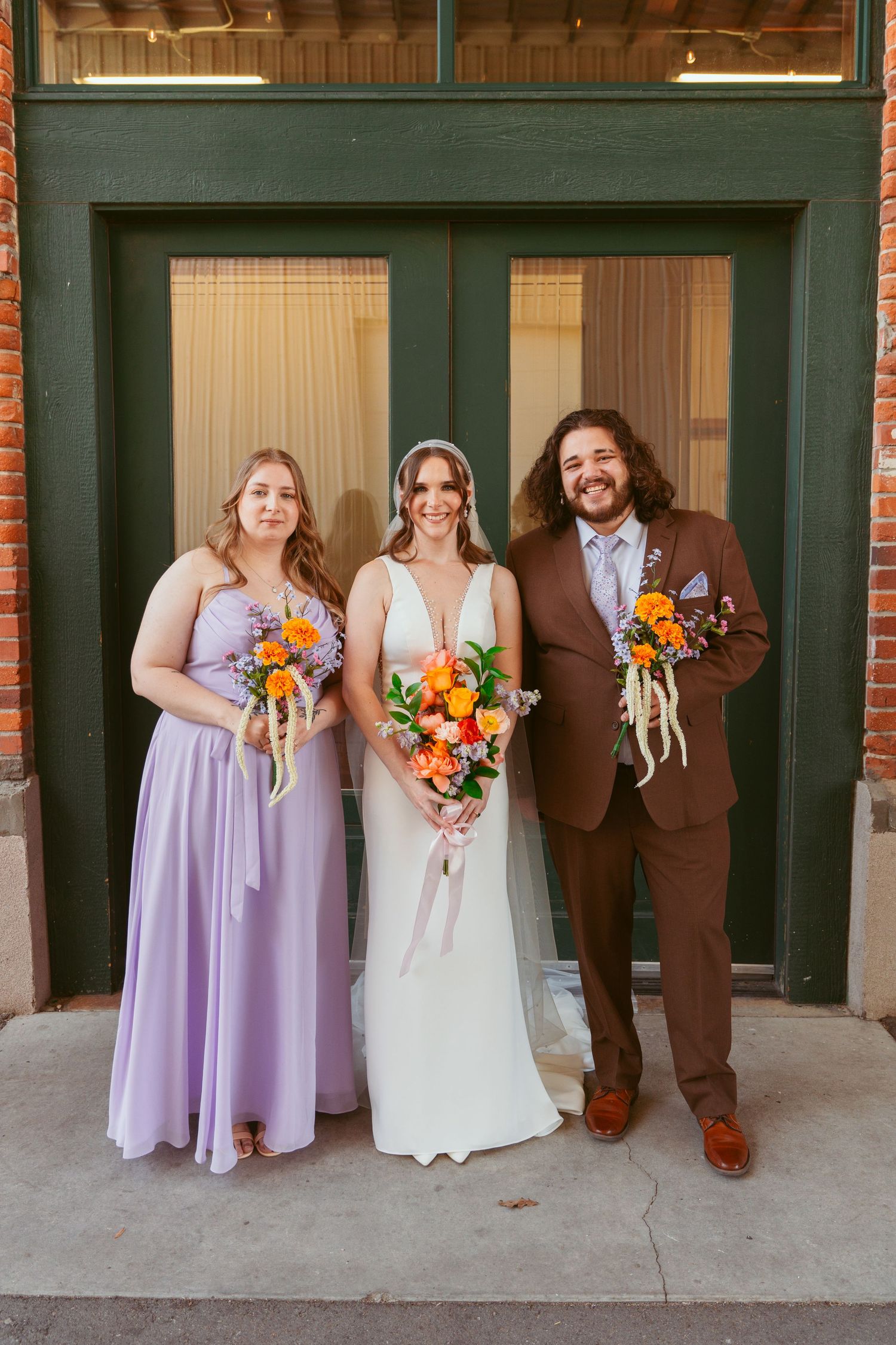 Wedding group portrait with lavender bridesmaid dress and brown suit against brick building.