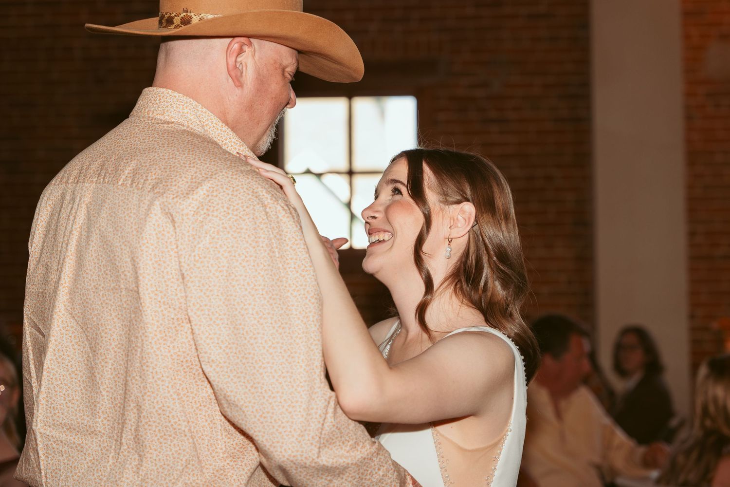 A couple performs their first dance at a wedding reception in a rustic brick-walled venue with string lights.