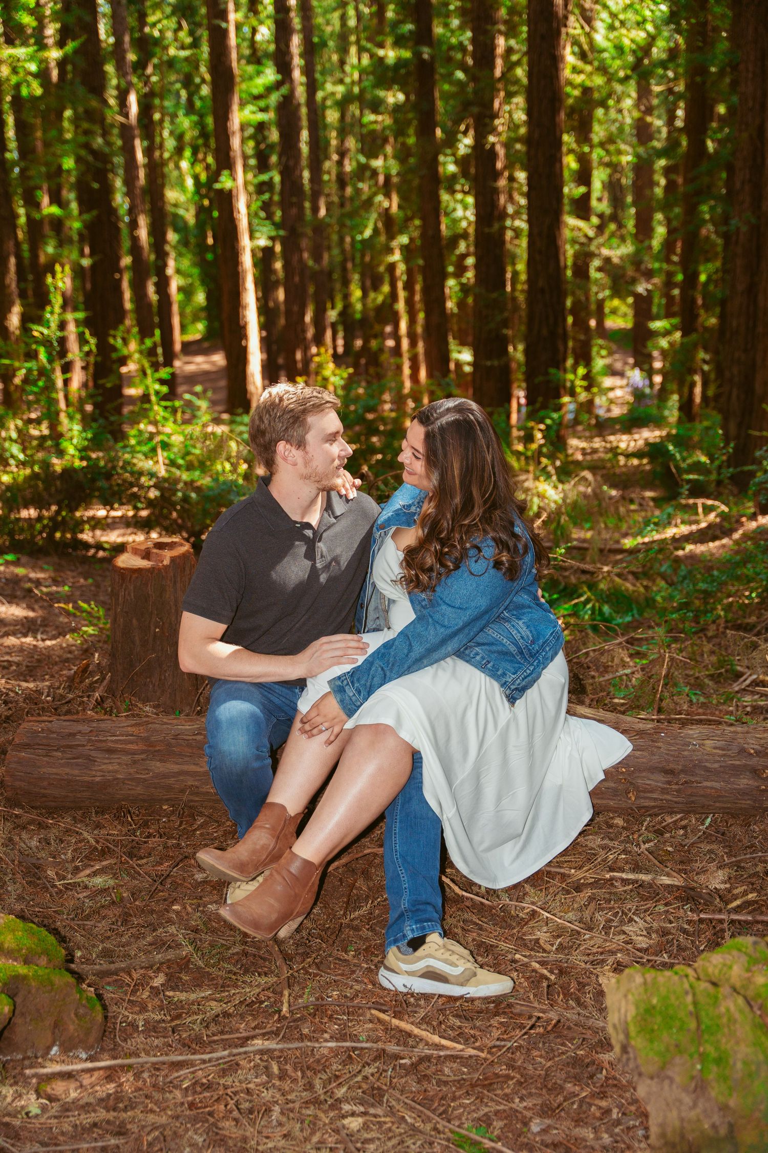 A playful couple sits together on forest ground surrounded by tall trees.
