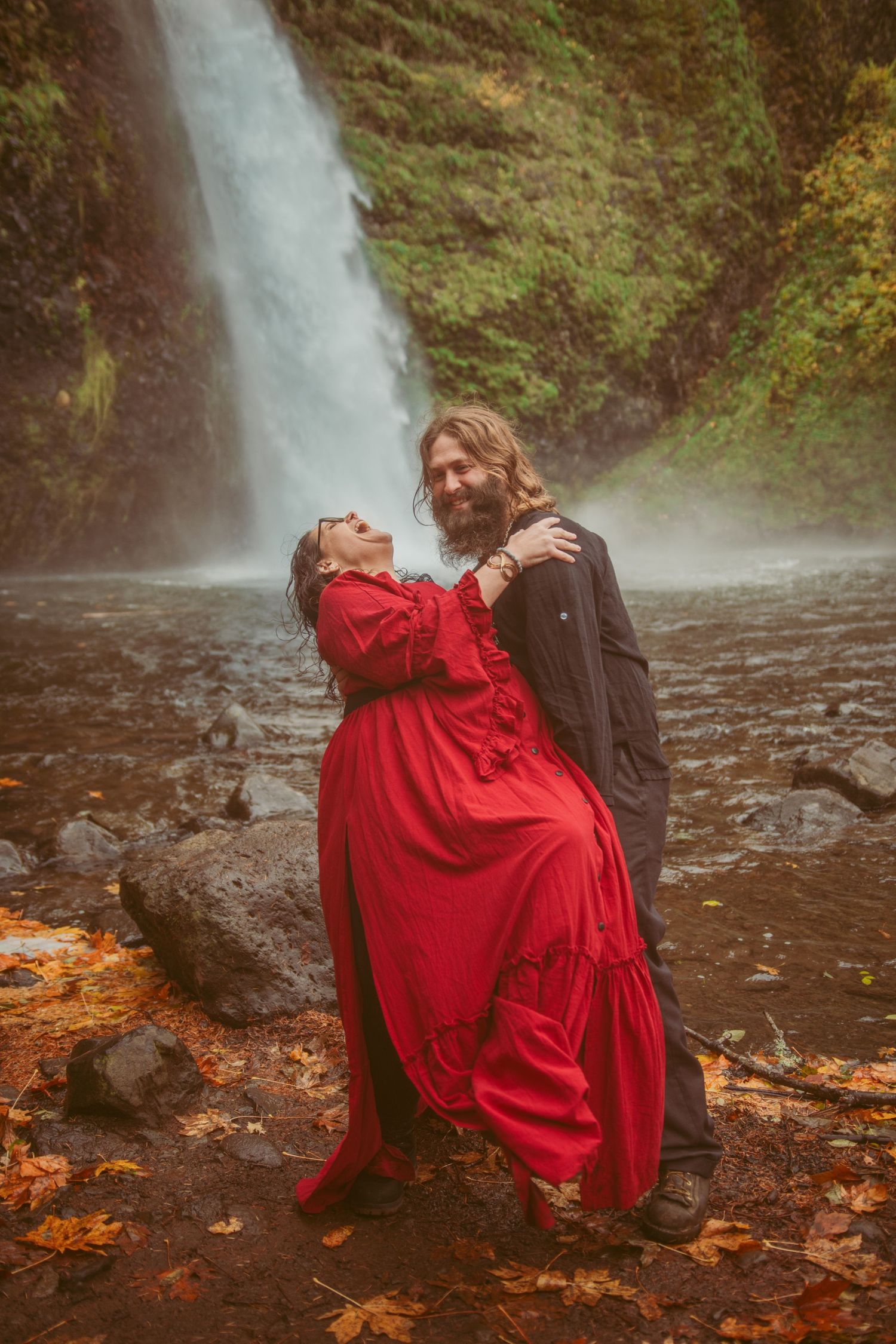 Romantic couple poses dramatically against misty waterfall with autumn leaves on ground.