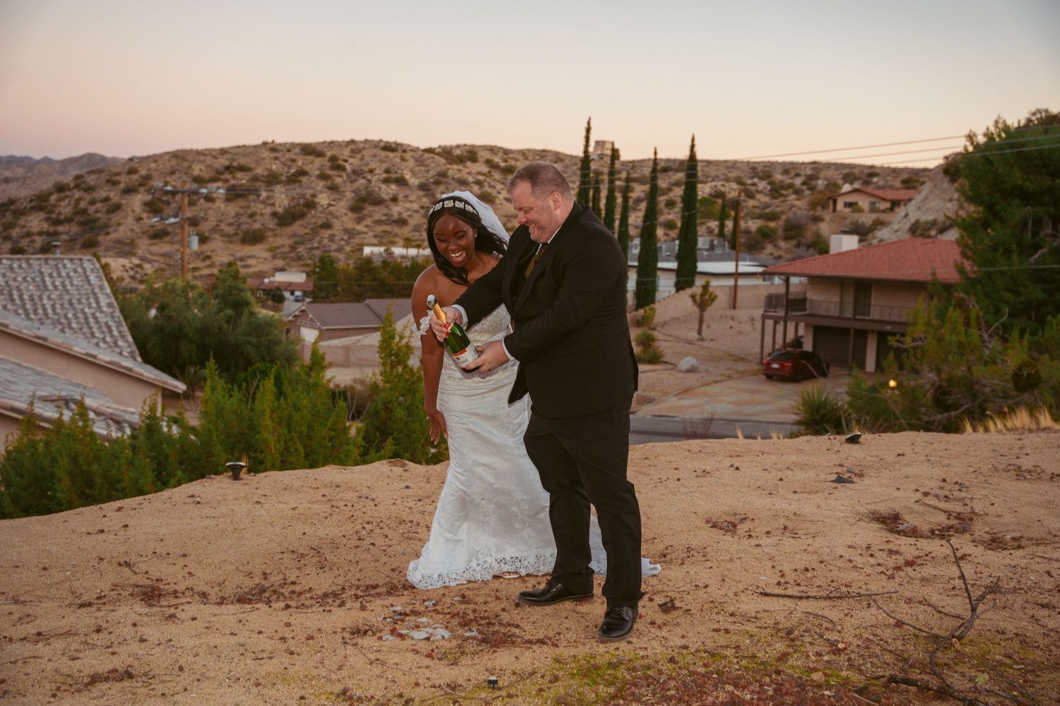 A couple in wedding attire poses against a desert landscape backdrop at sunset.