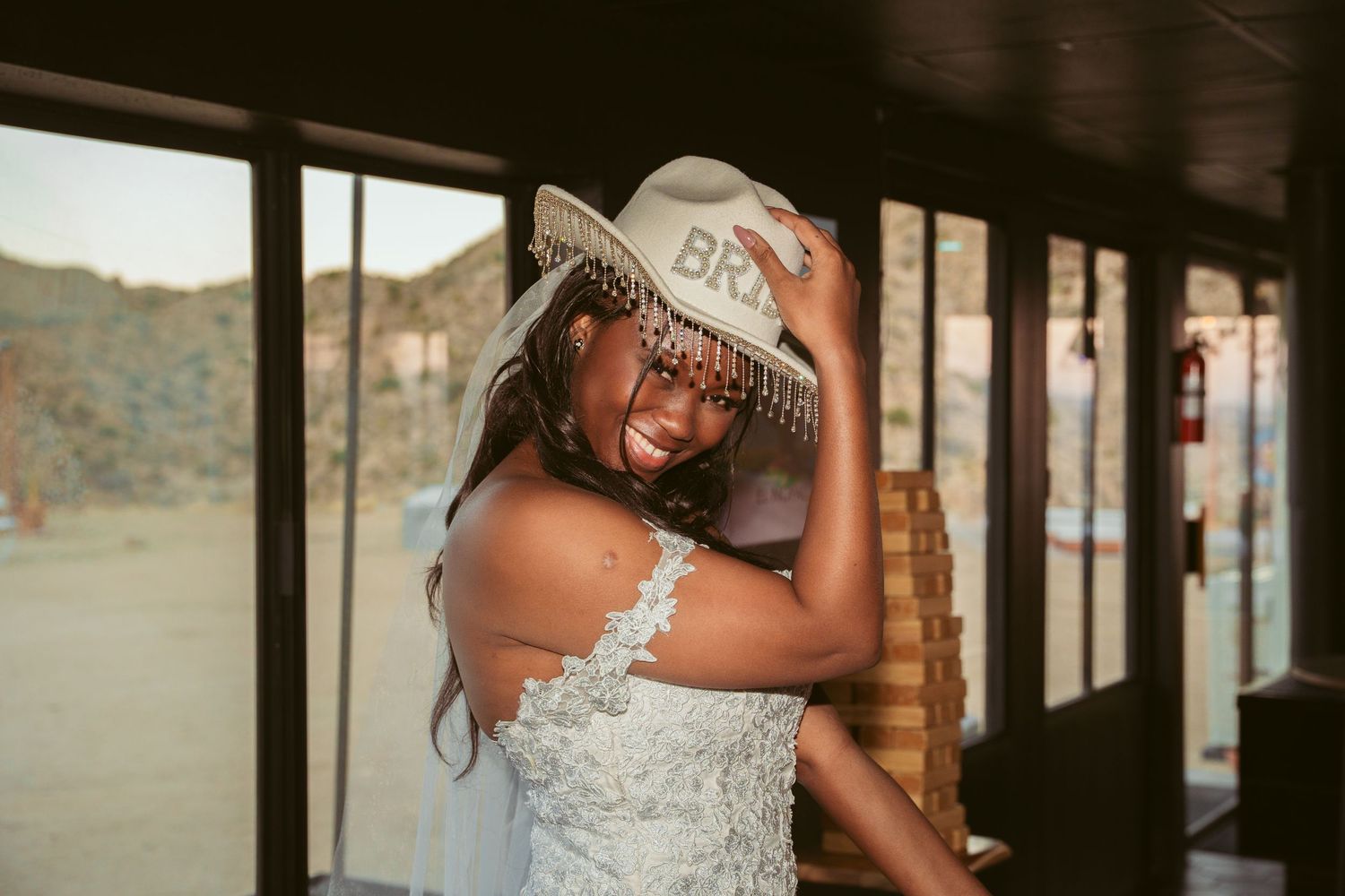 Bride in a white lace dress poses near window light.