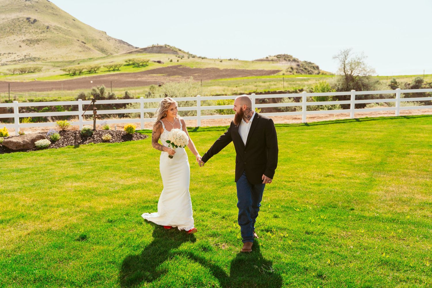 Couple walks hand in hand across green field with mountain landscape behind them.