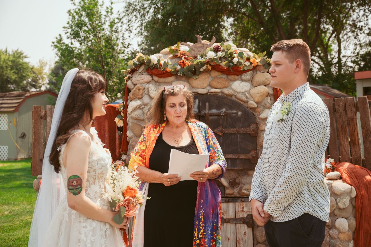 Wedding party gathers for ceremony in front of decorative stone pillars.