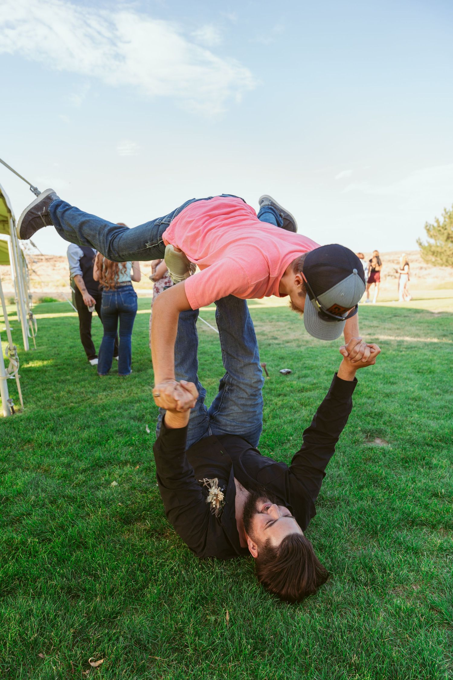 Playful guests perform acrobatic moves on the grass during an outdoor wedding celebration.