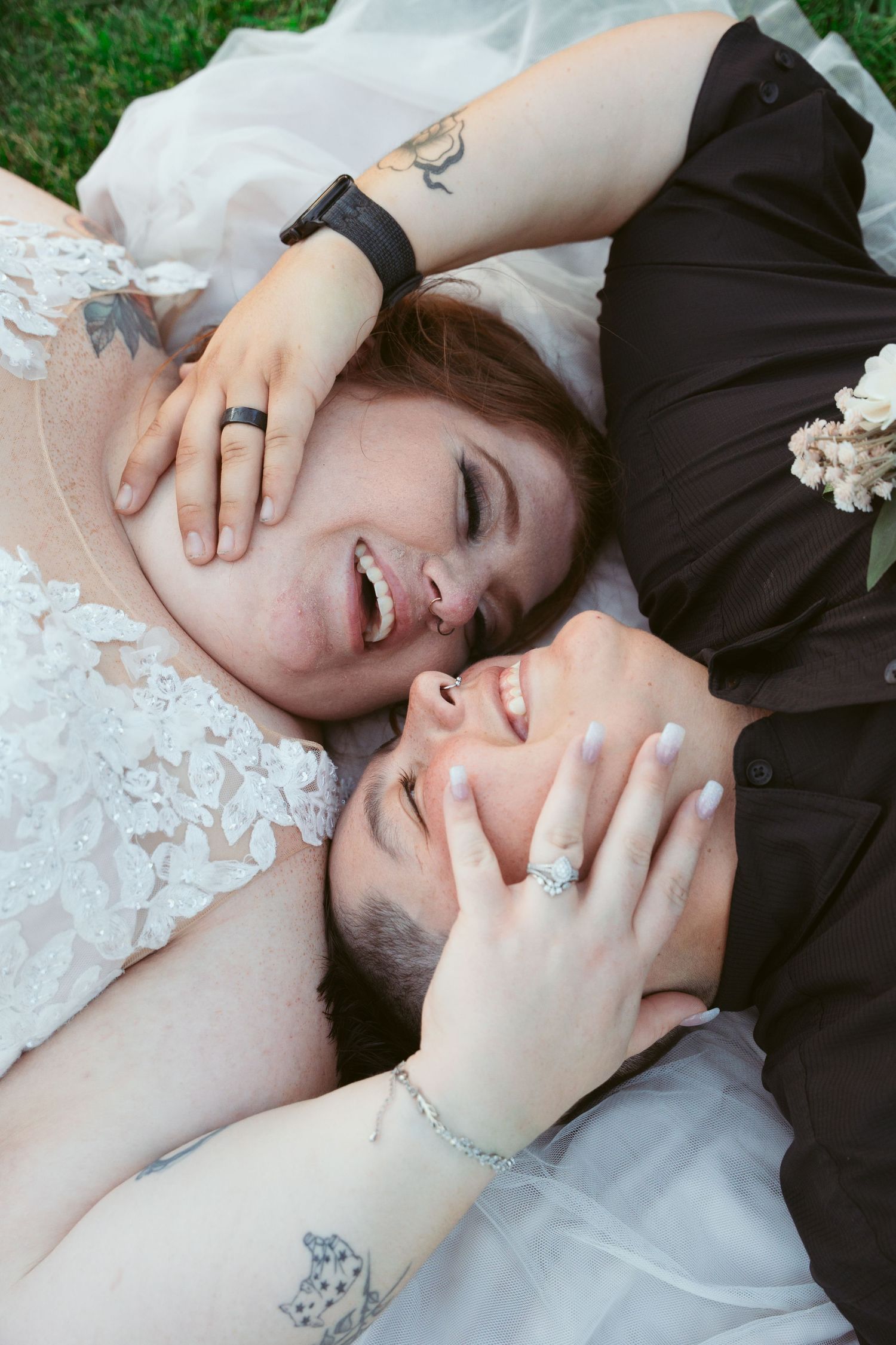 A couple lying on grass laughing together in their wedding attire with visible tattoos.