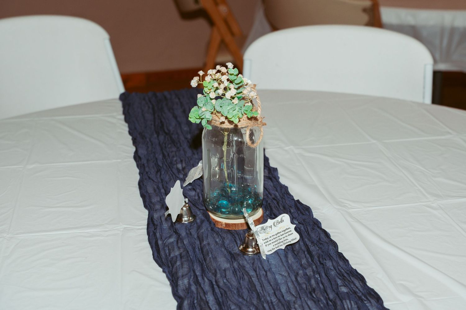 Wedding reception table setup with white linens, navy blue runner, and decorative cake display.