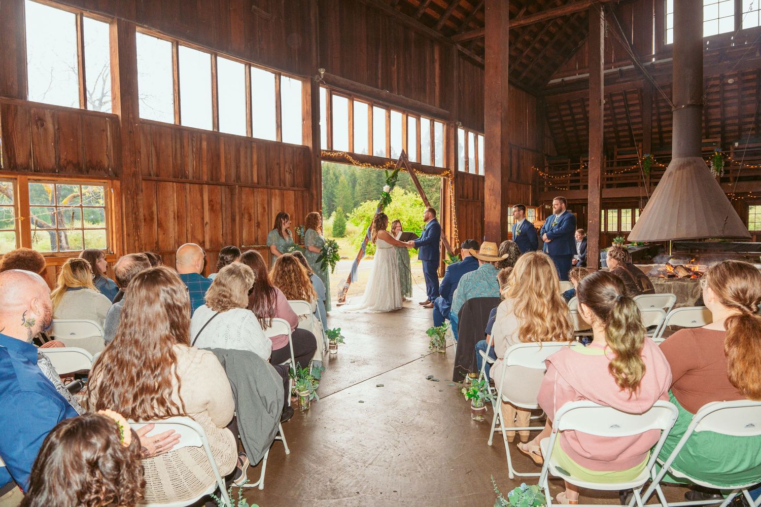 Wedding ceremony guests seated in rustic wooden barn venue with string lights overhead.