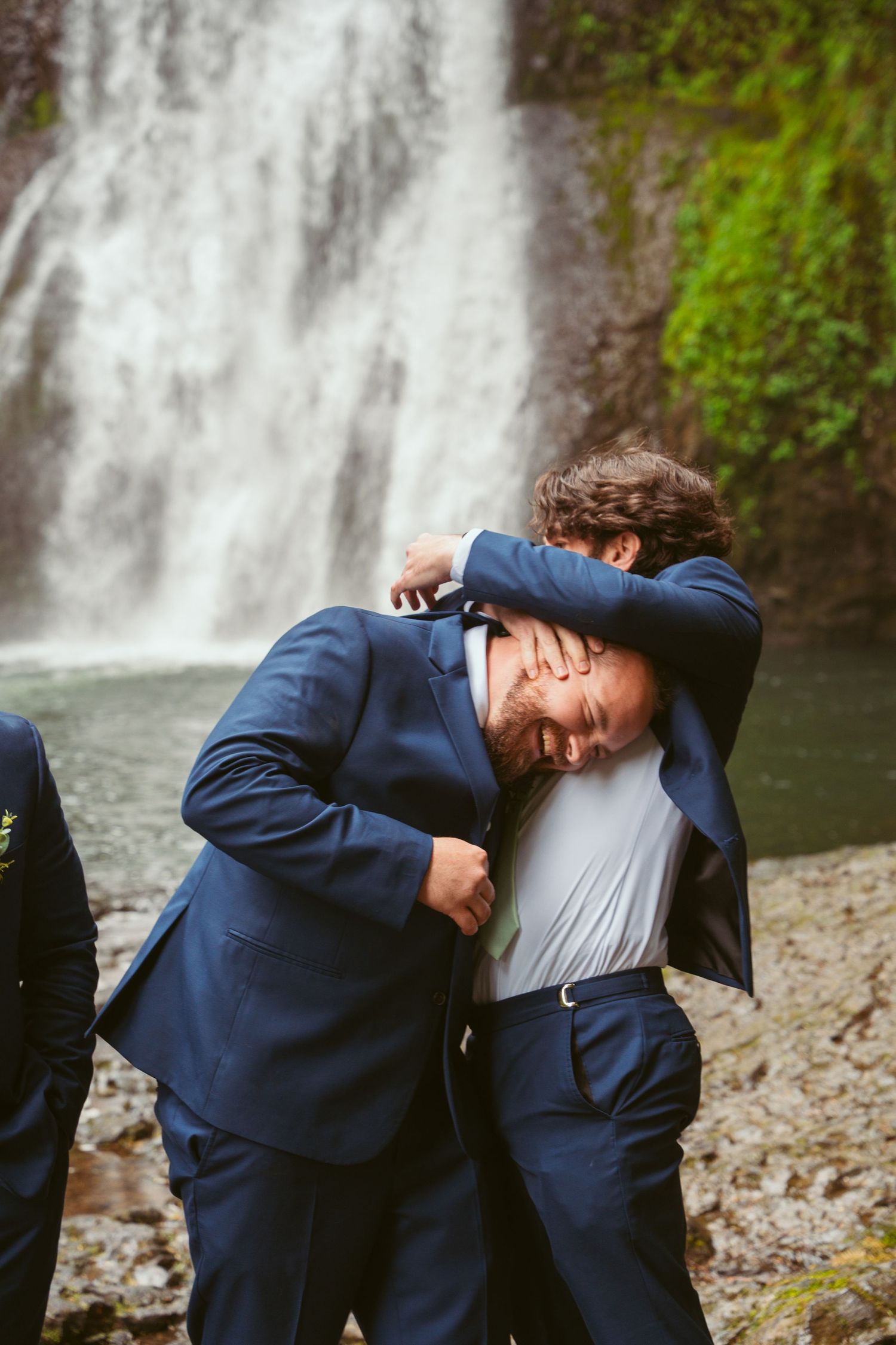 Groomsmen in navy suits playfully pose by a cascading waterfall.