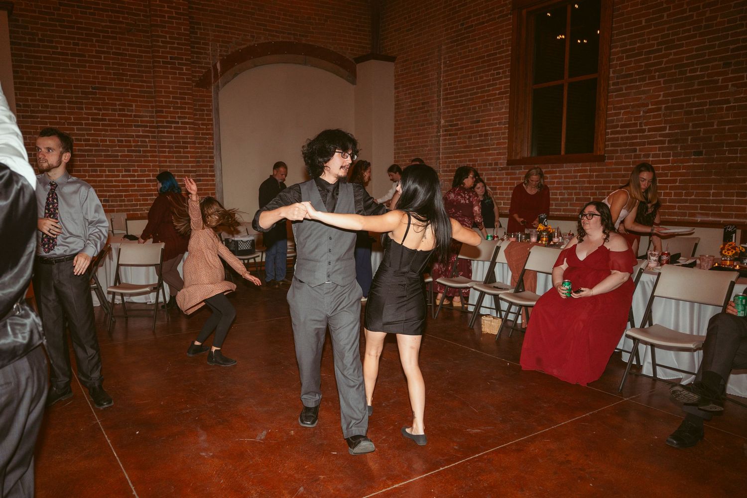 Guests enjoying swing dance moves at a reception in an industrial brick venue with string lights.