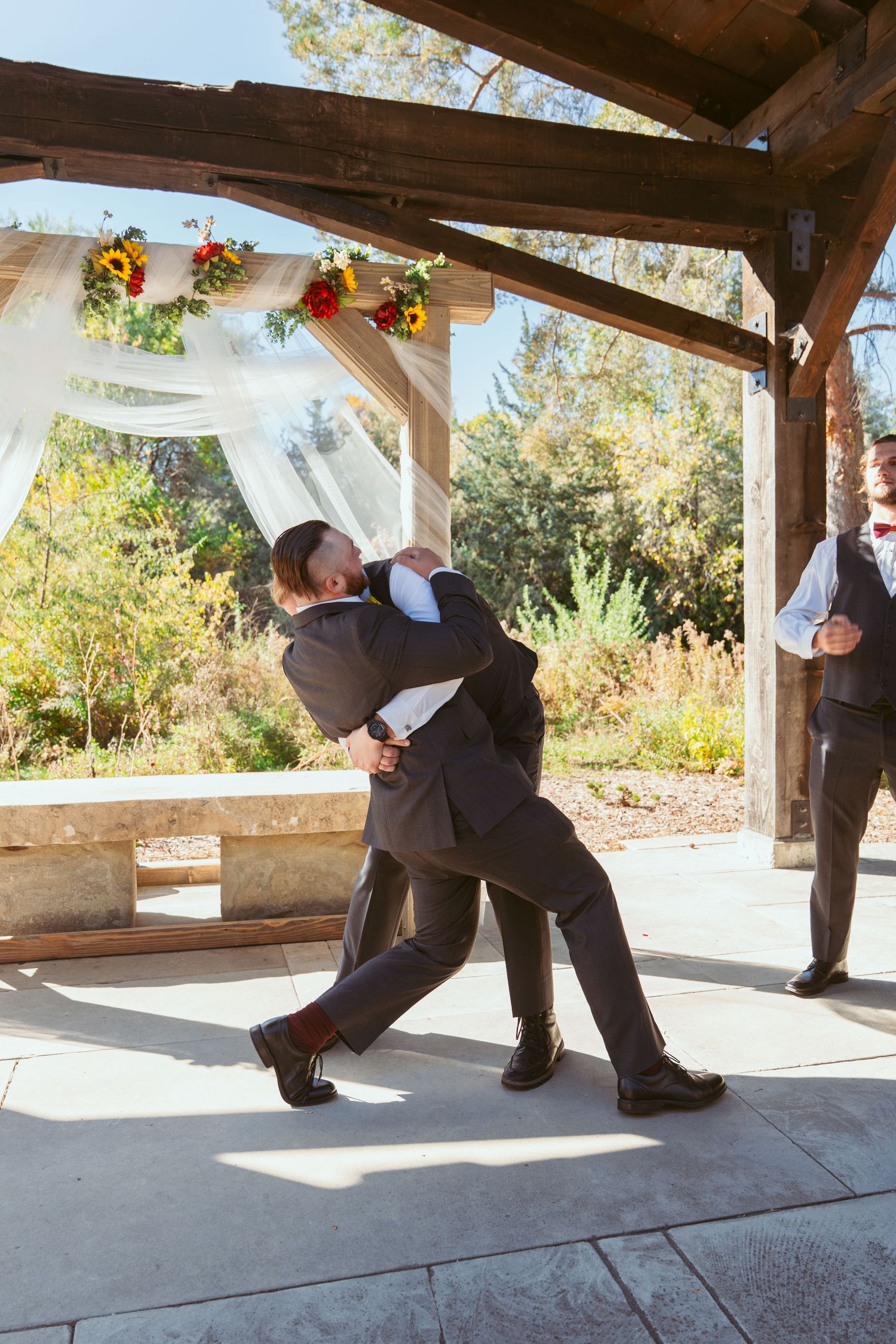 Wedding party members perform a fun falling down sequence in formal attire under a covered outdoor venue.