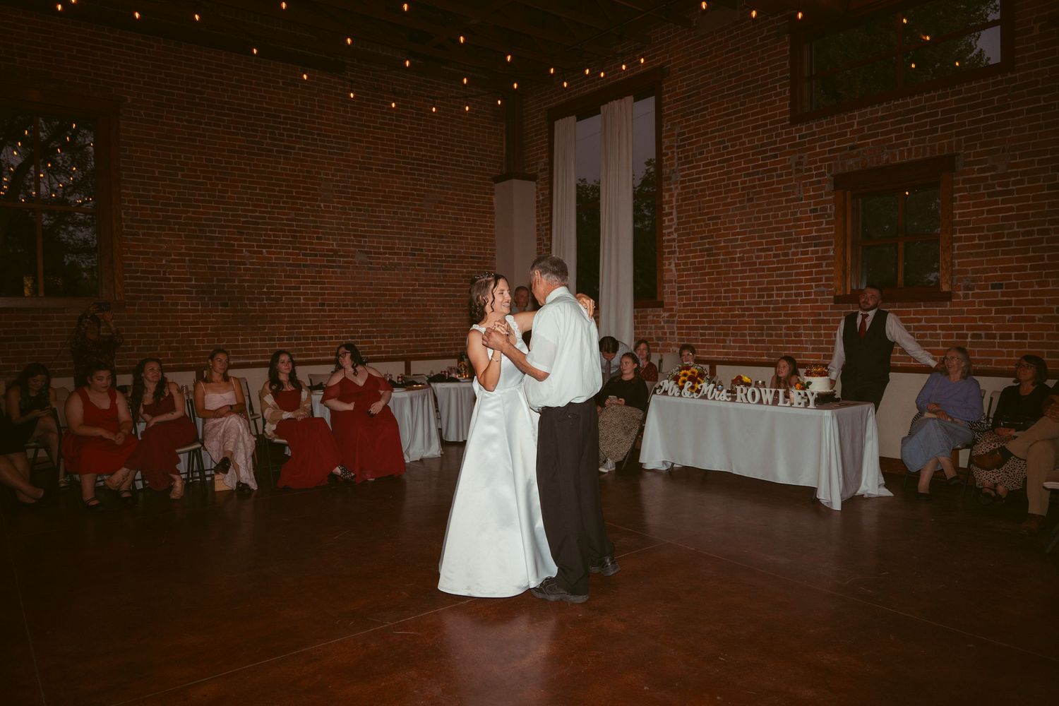 Wedding guests watch a couple dancing in a spacious brick-walled reception venue.