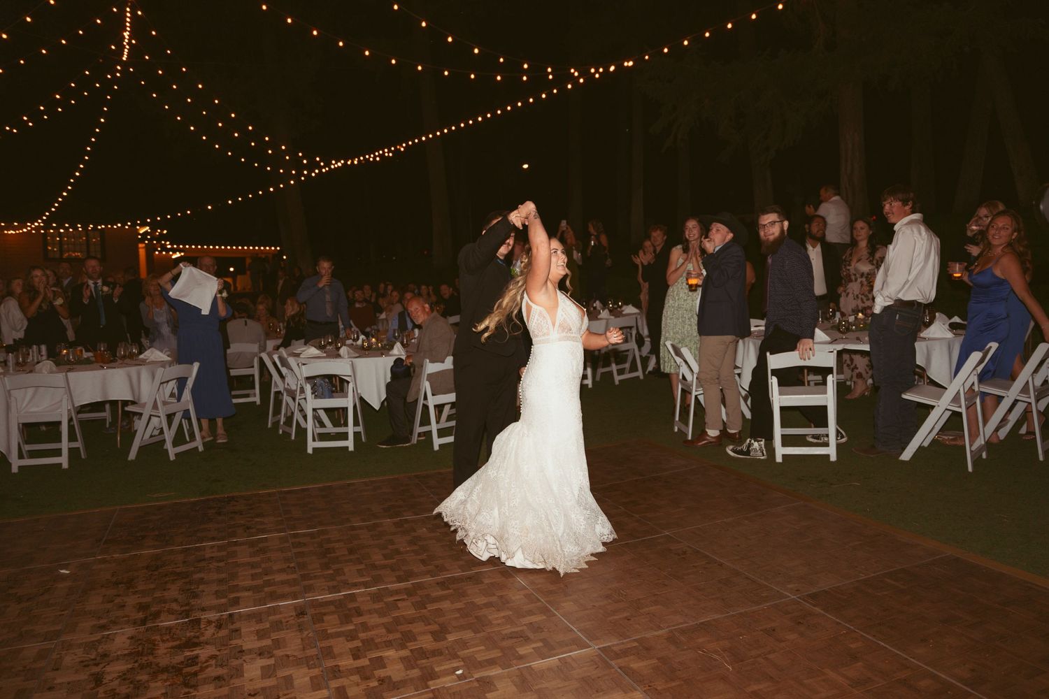 Outdoor wedding reception with guests seated at tables and a dance floor under string lighting.