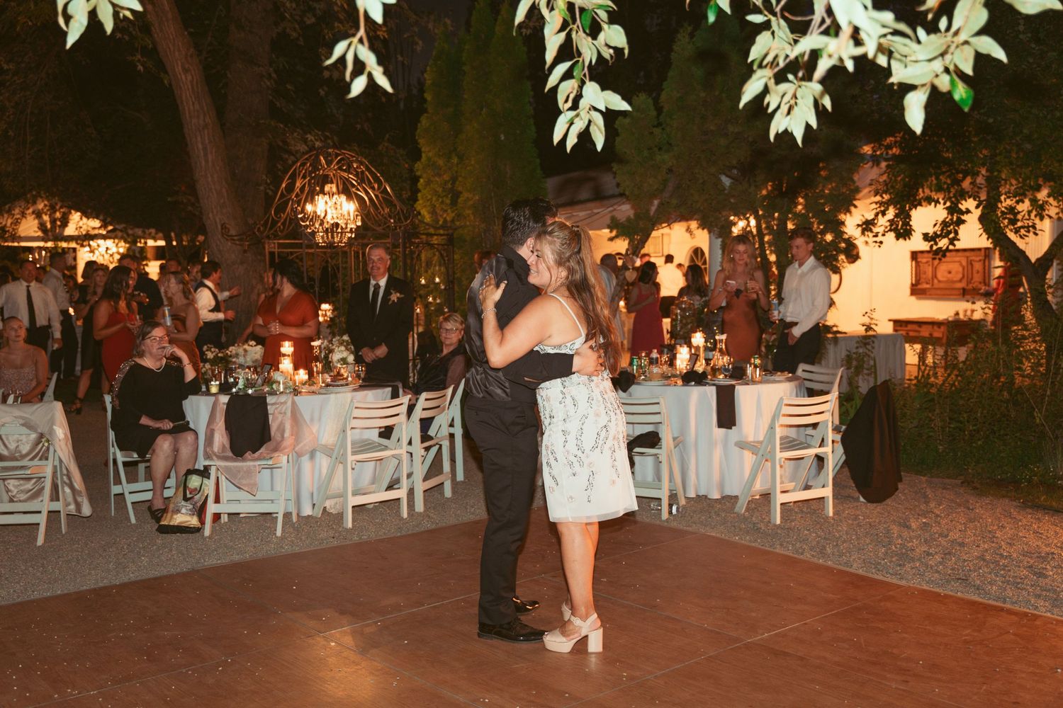 Outdoor wedding reception venue with white chairs and decorative lighting under trees.
