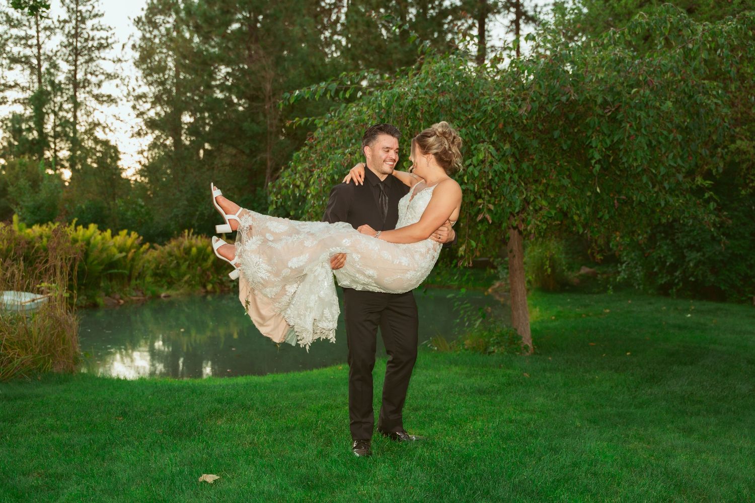 Groom carries bride in flowing dress during sunset photoshoot by pond.
