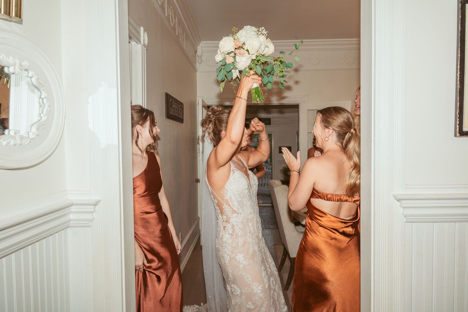 Wedding party members in copper-colored dresses celebrate in an indoor hallway.