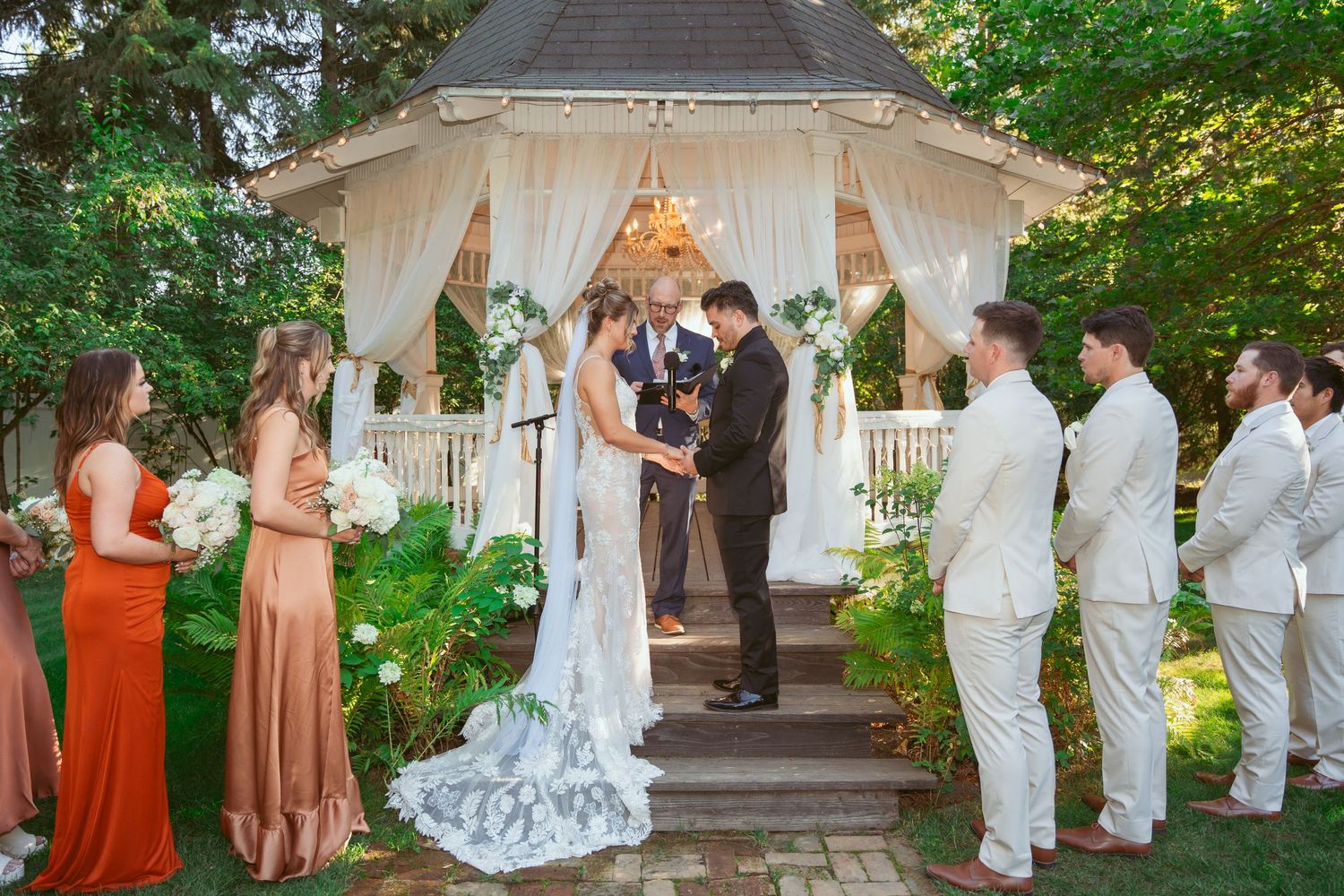 Outdoor wedding ceremony with white gazebo, guests in formal attire, and rustic decor.