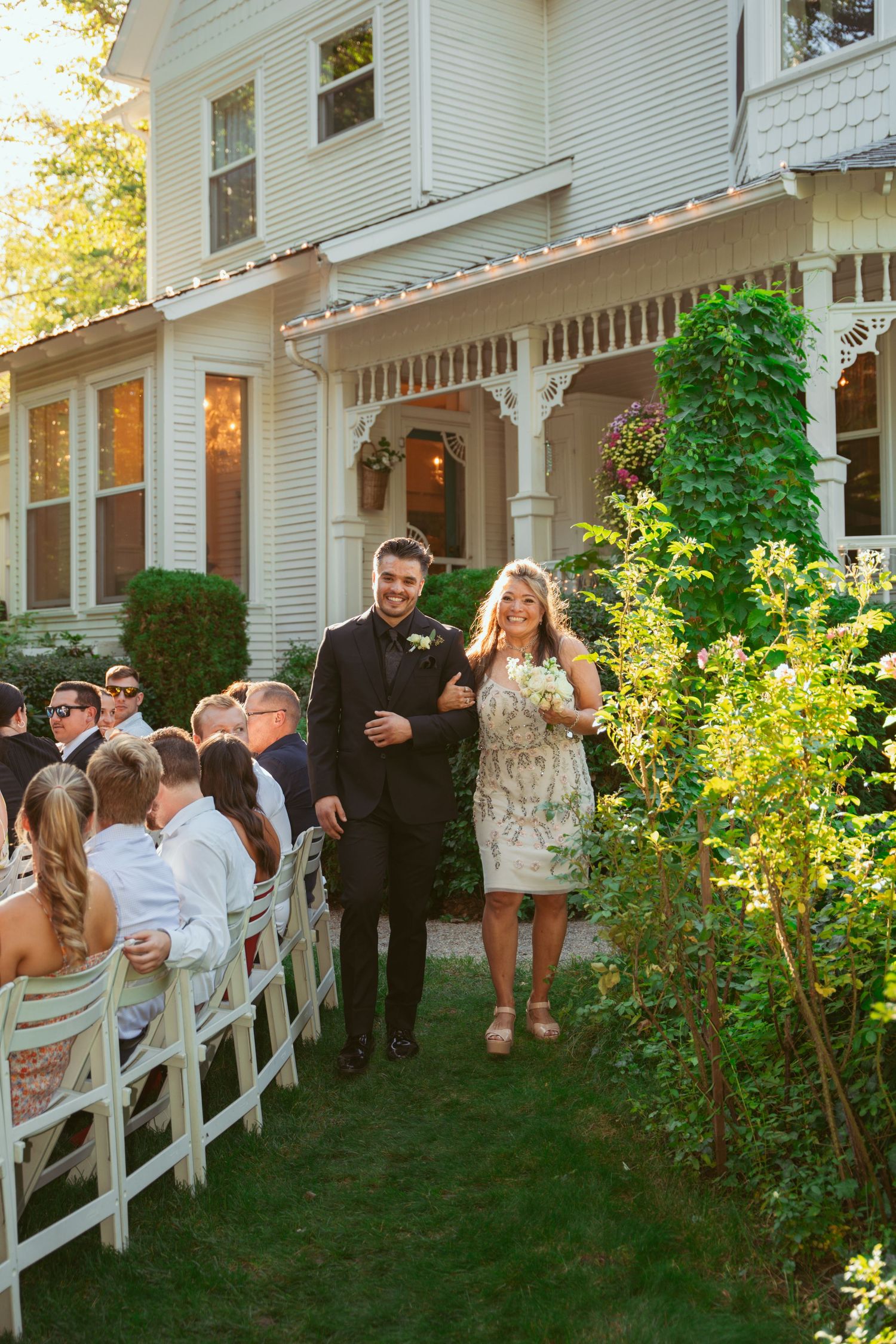Wedding guests enjoy a casual outdoor gathering along a white fence at sunset during a backyard reception.