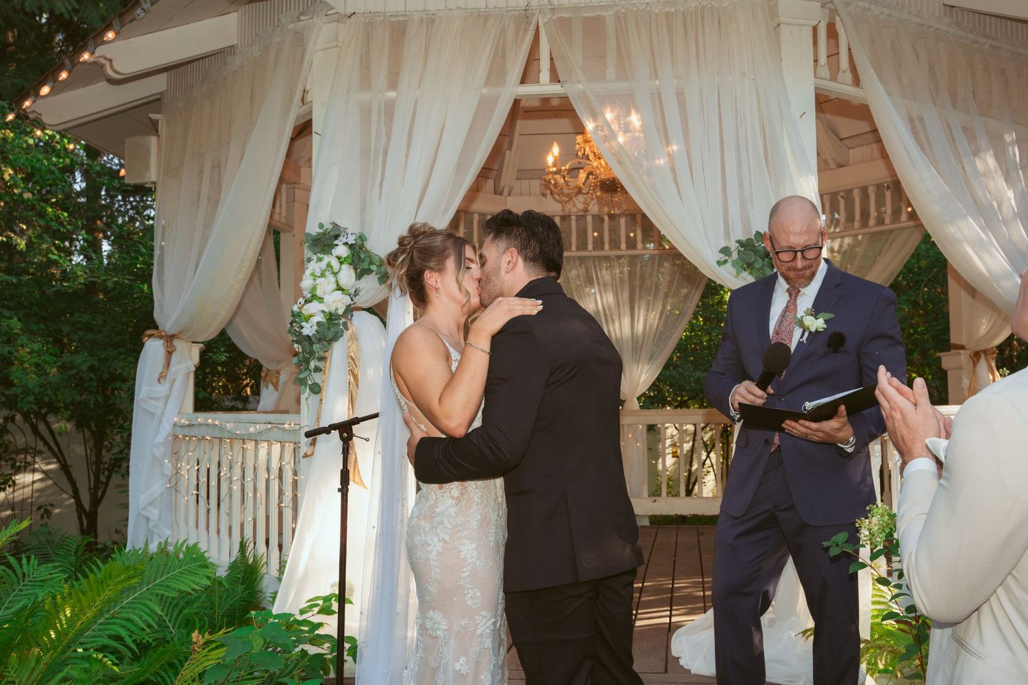 Romantic wedding ceremony takes place under a draped gazebo with chandelier and white curtains.