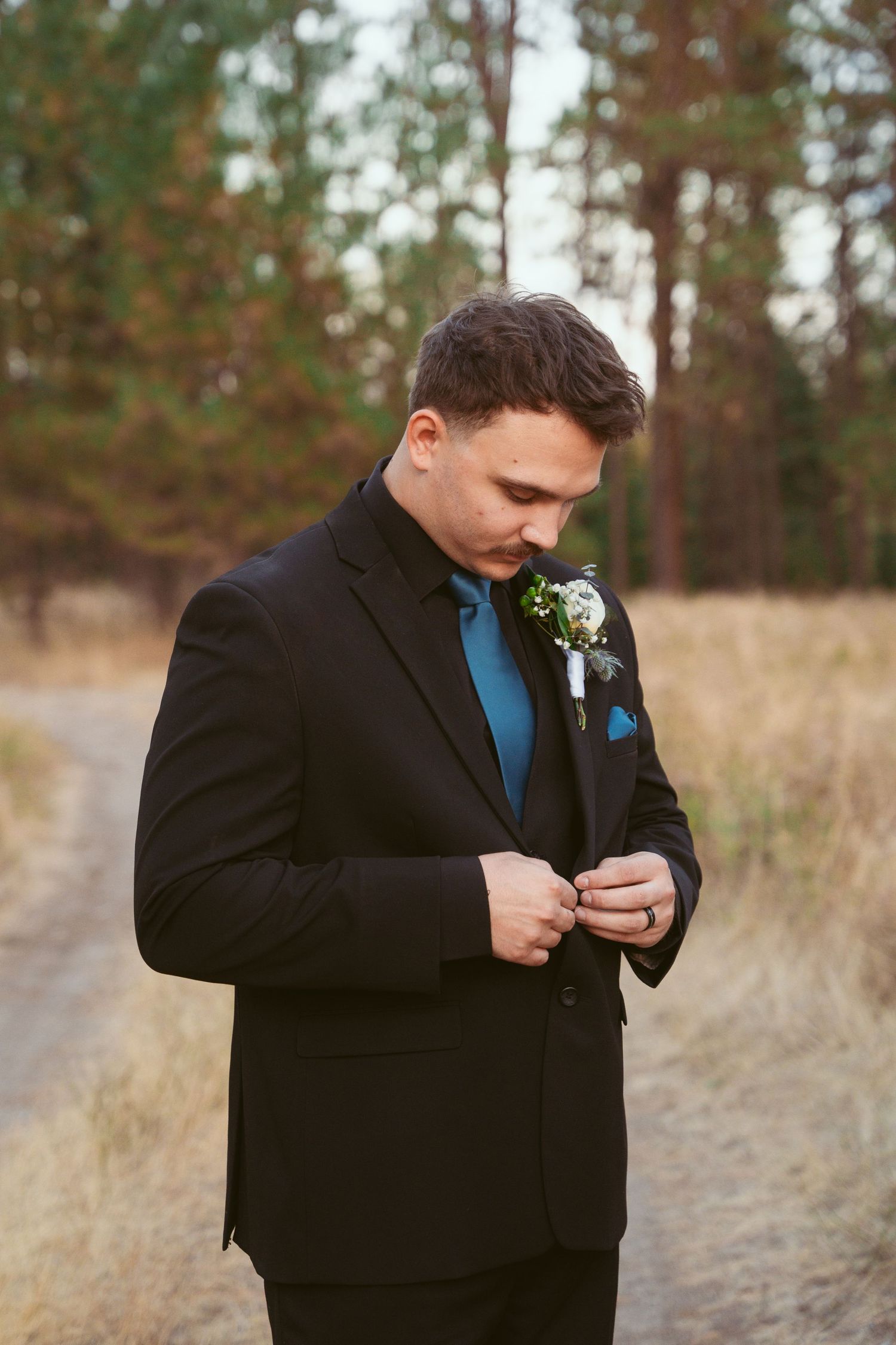 Groom in black suit with blue tie adjusts jacket in rustic outdoor setting.